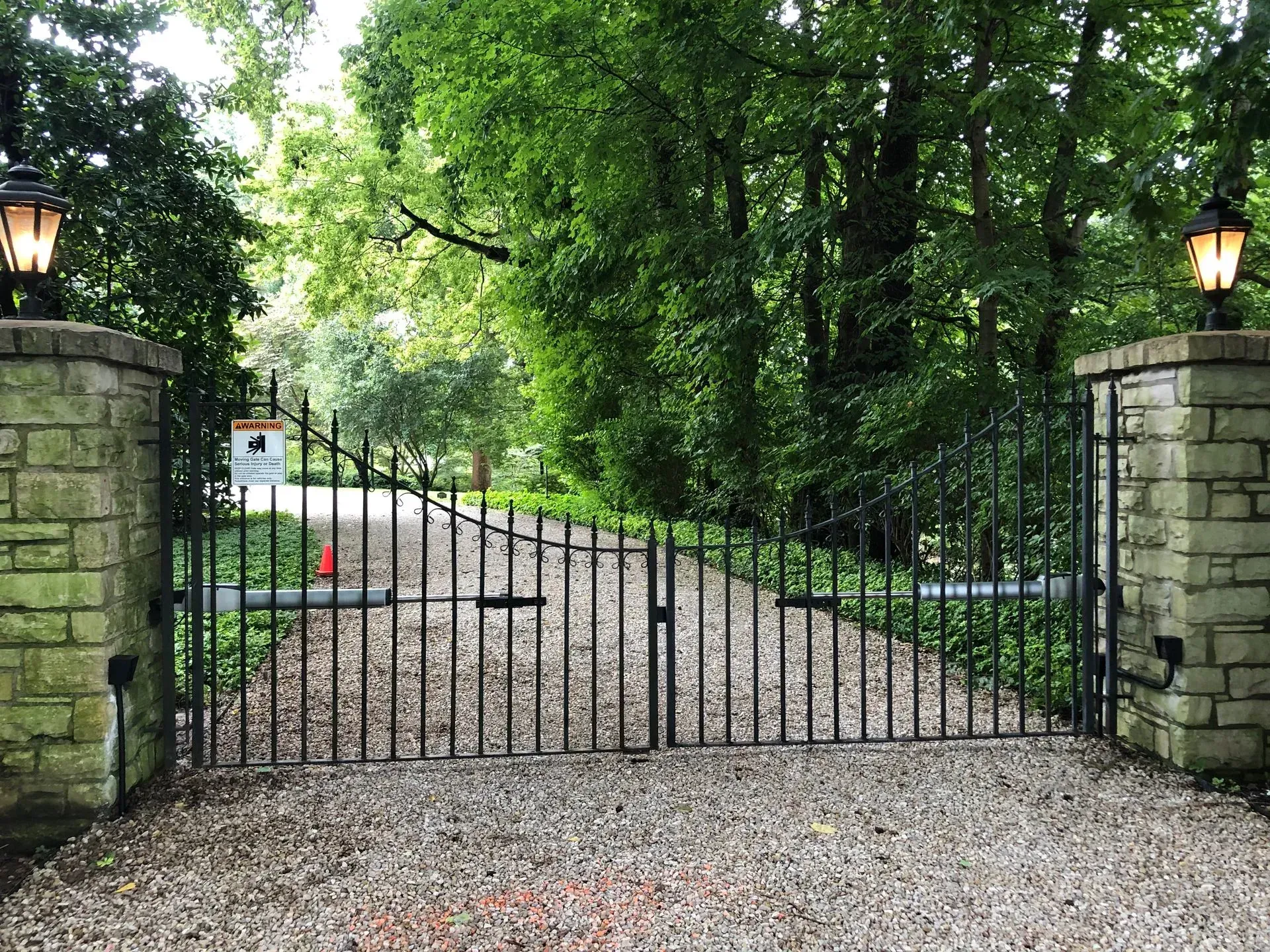 A wrought iron gate is open to a gravel driveway surrounded by trees.