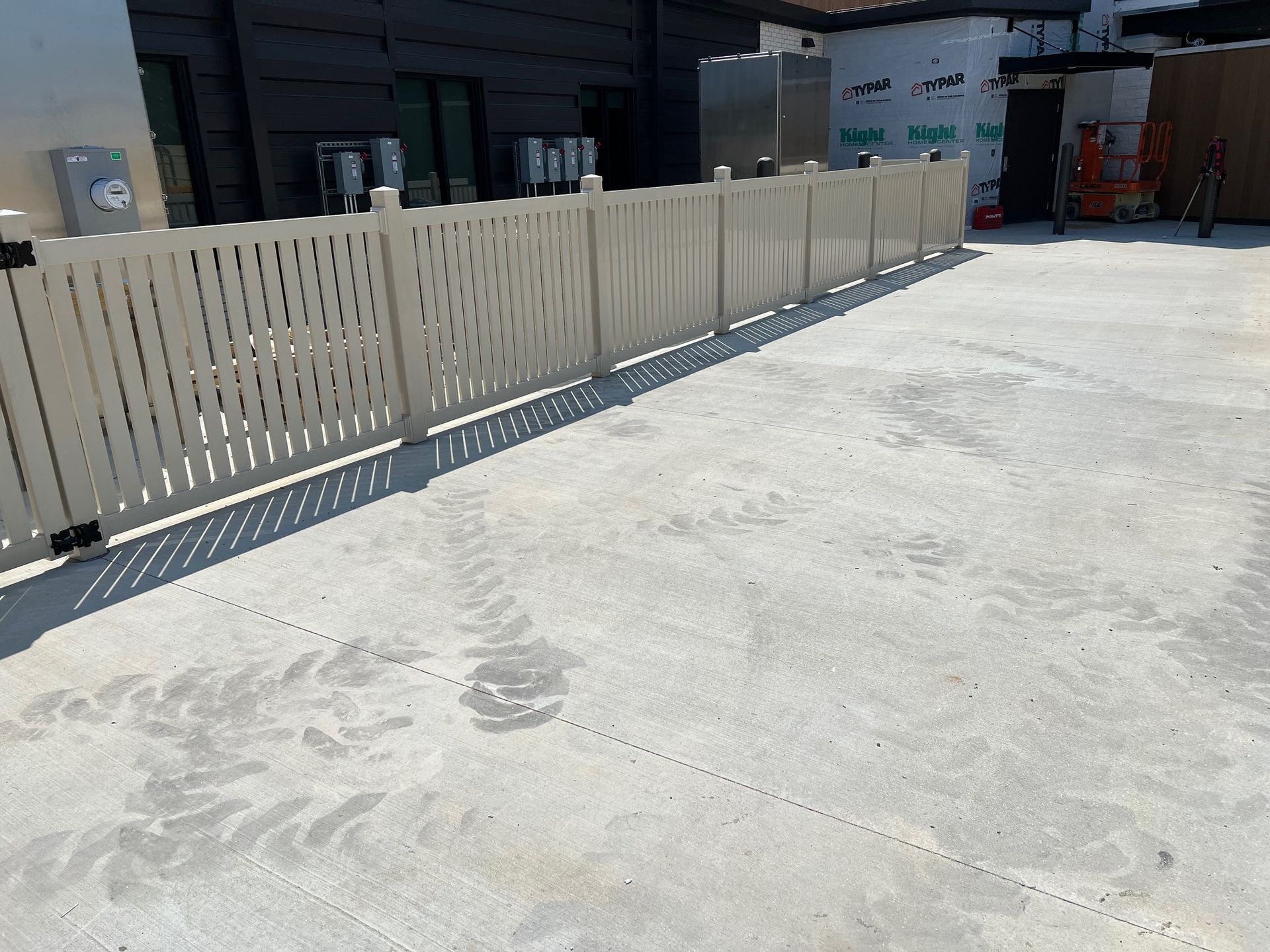 A white fence surrounds a concrete driveway in front of a building under construction.