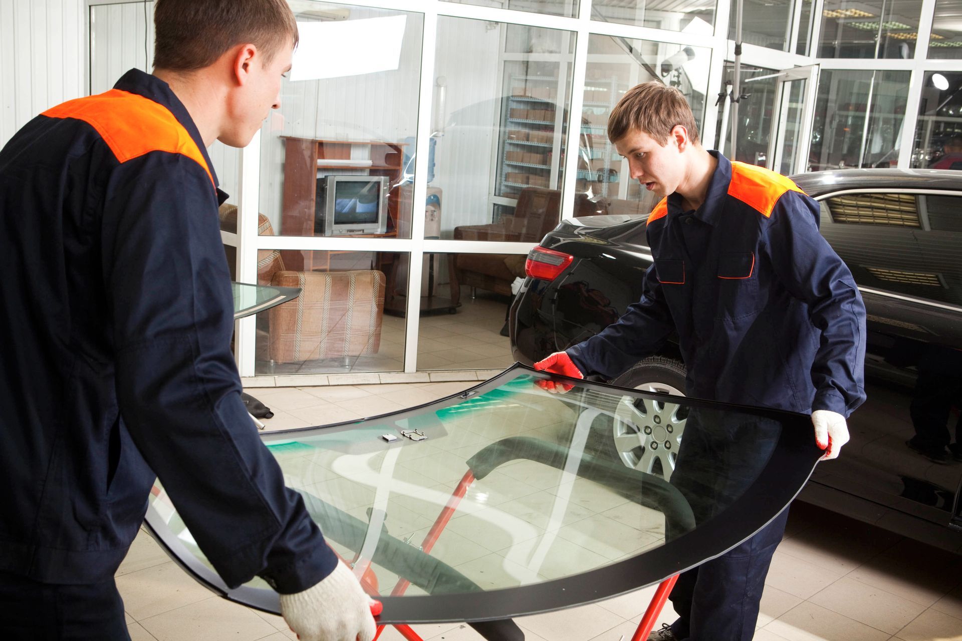 Two auto technicians in uniforms carefully carrying a replacement windshield inside a repair shop.