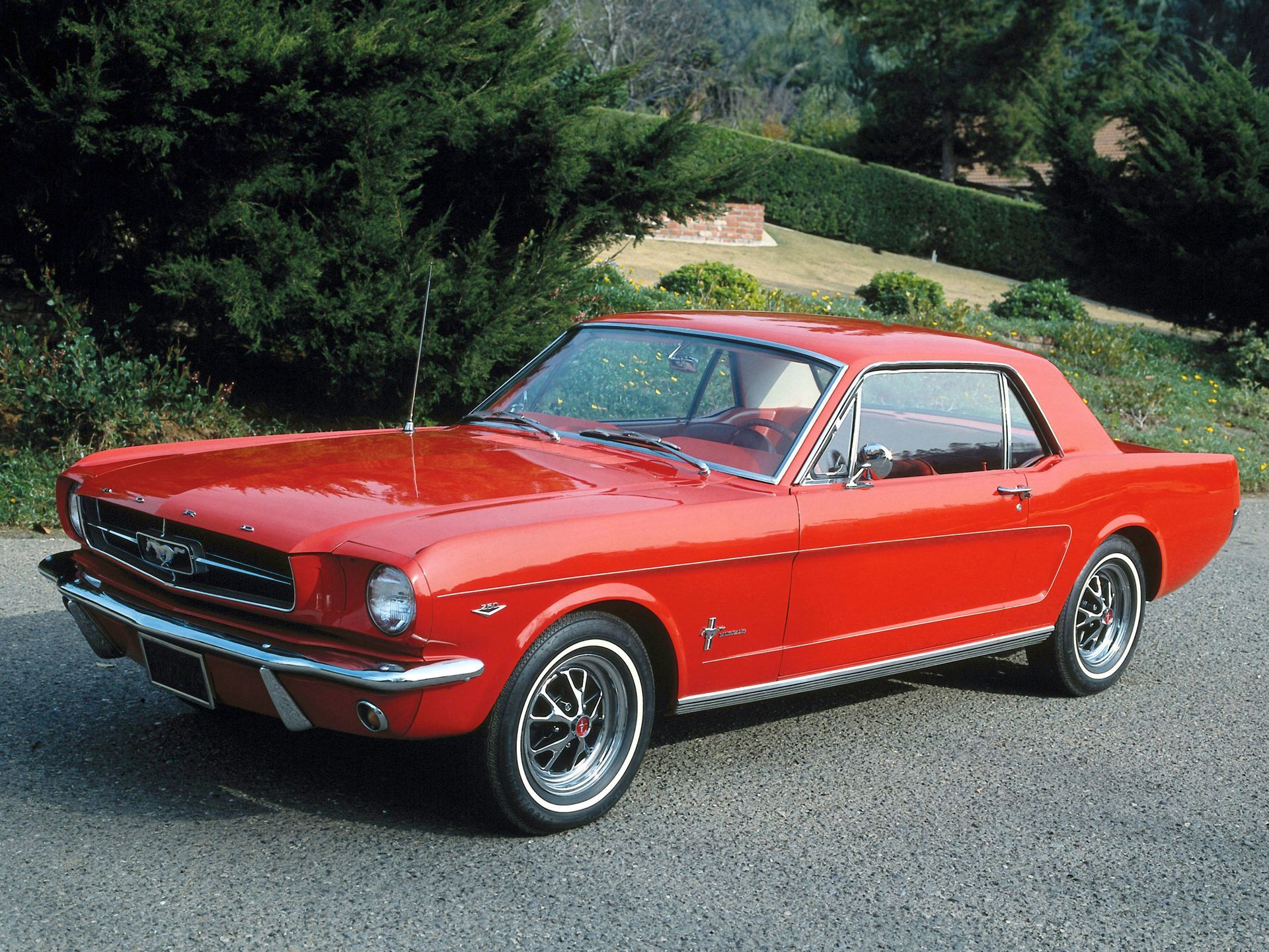 Bright red vintage Ford Mustang parked outdoors on a road with trees and bushes in the background.