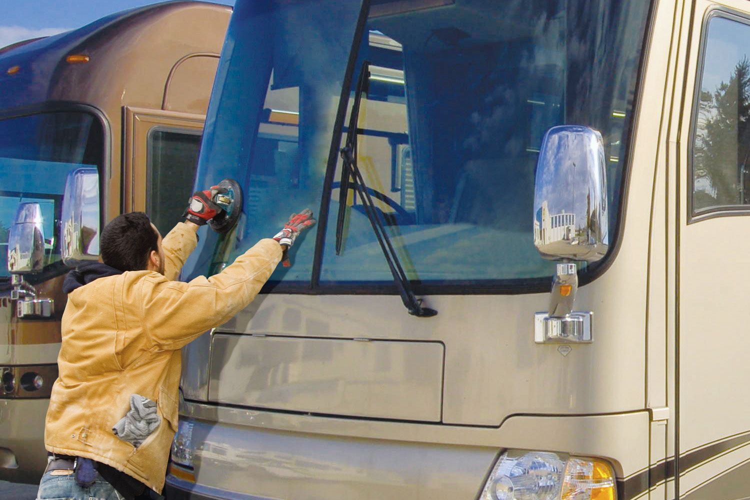 Worker in a tan jacket installing a large windshield on a motorhome using a suction tool.