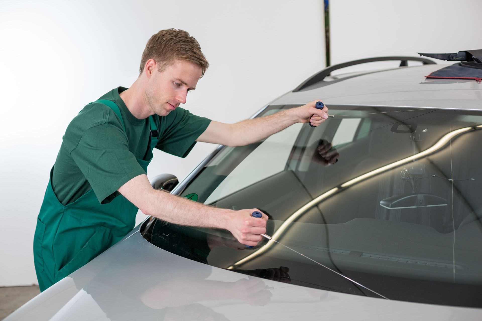 A mechanic in green overalls installing a new windshield on a silver car using suction tools.