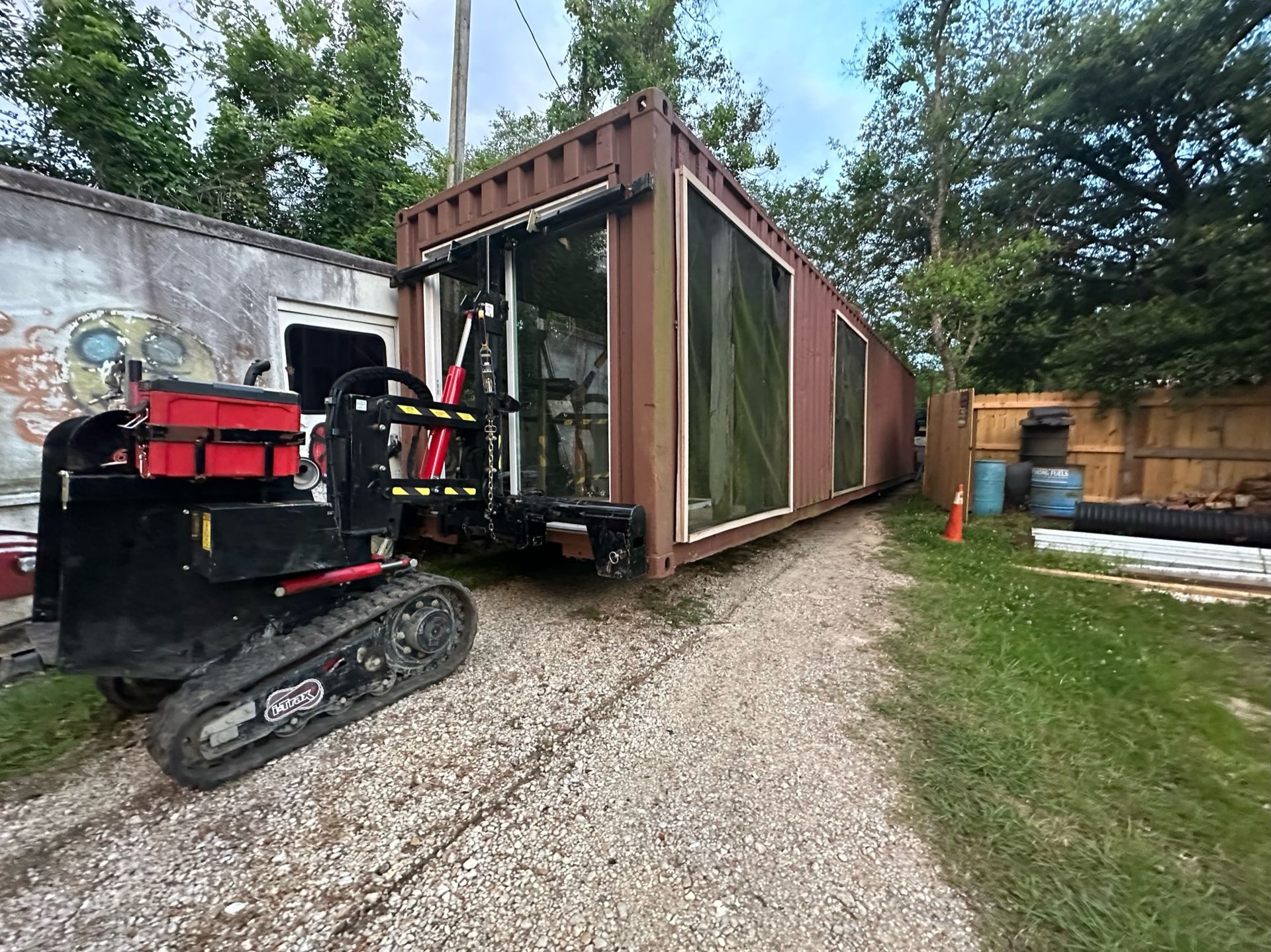 A tracked carrier vehicle moves a brown shipping container with glass window cutouts through a gravel yard.