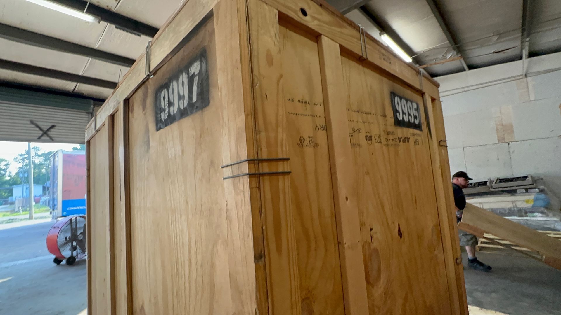 A large wooden shipping crate marked with the numbers 9997 and 9995 stands inside a warehouse with a person in the background.