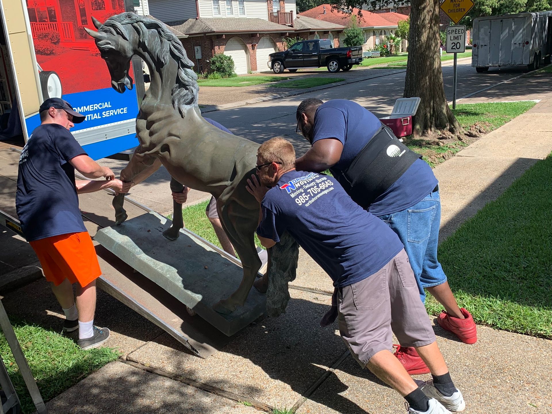Three movers in blue shirts guide a large, bronze-colored horse statue onto a ramp to load it into a moving truck.