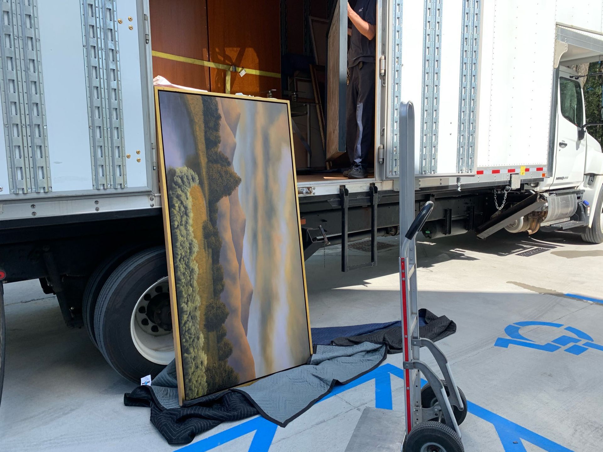 A person unloads a large landscape painting from a moving truck into a parking space.