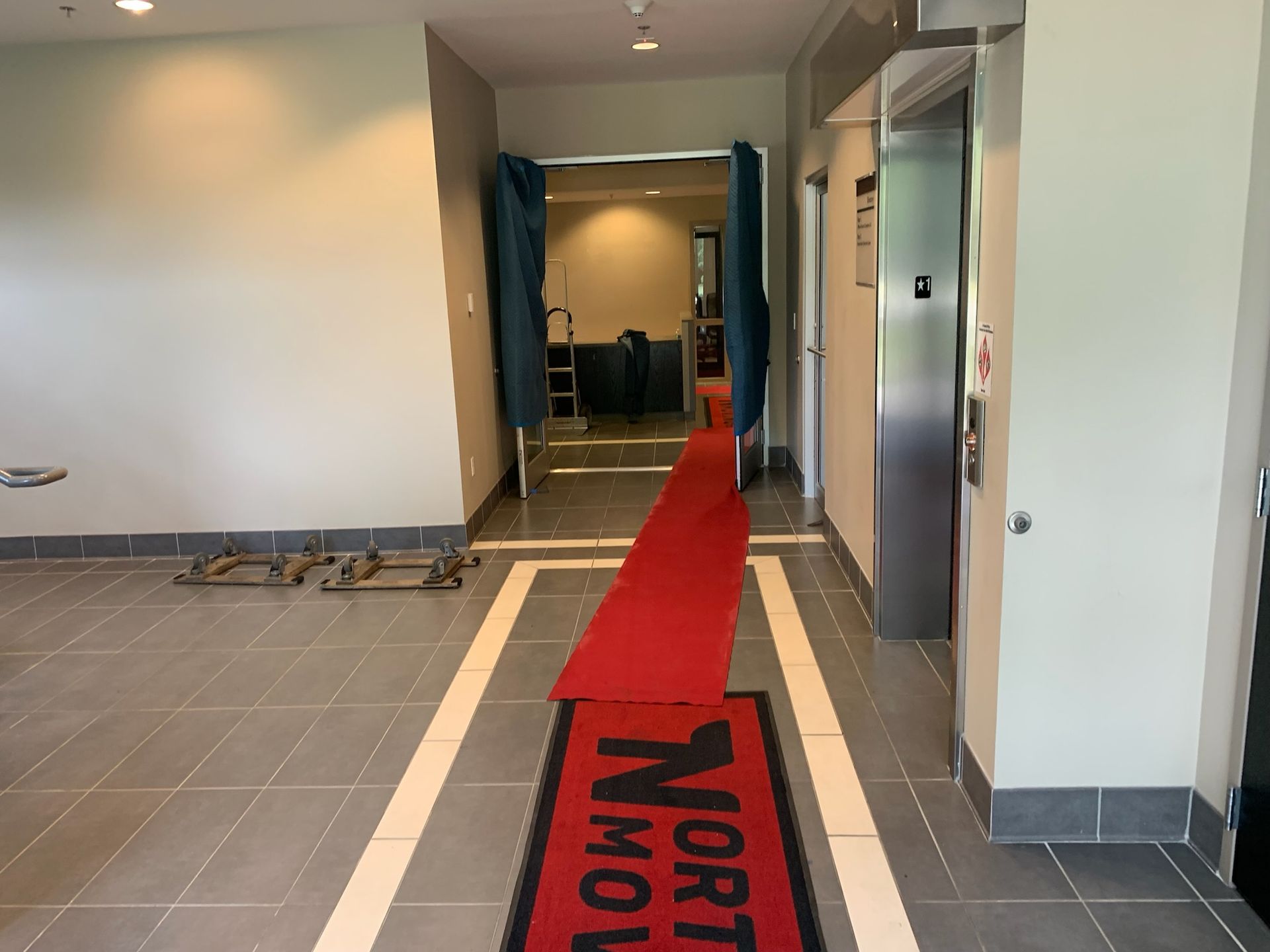 A red runner leading to an open doorway with blue curtains and an elevator to the right in a modern hallway.