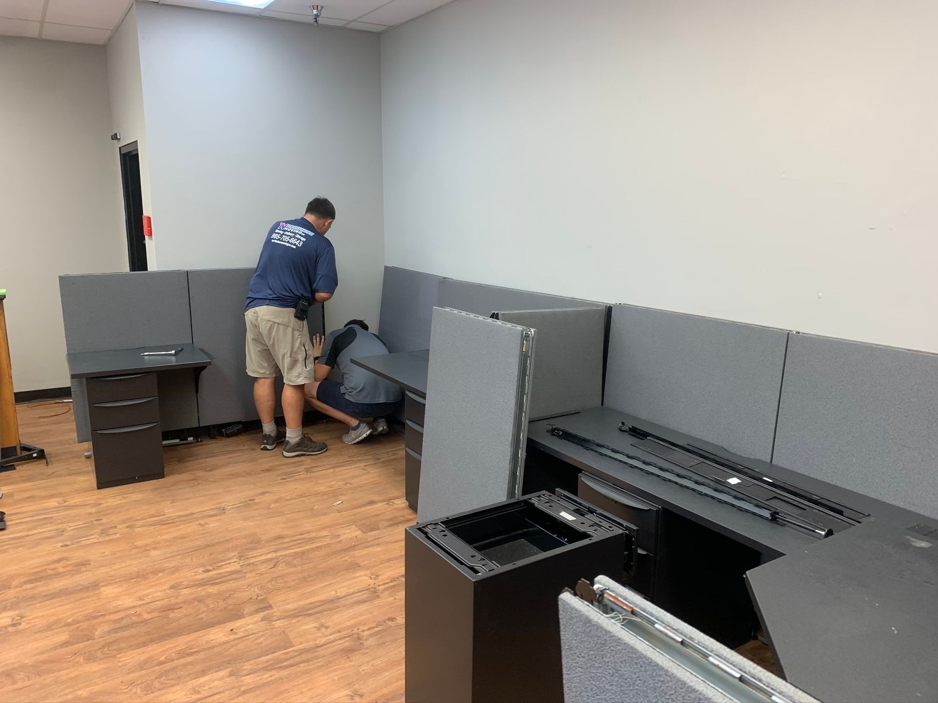 Two people assemble gray office cubicles in an empty room with wood-look flooring.