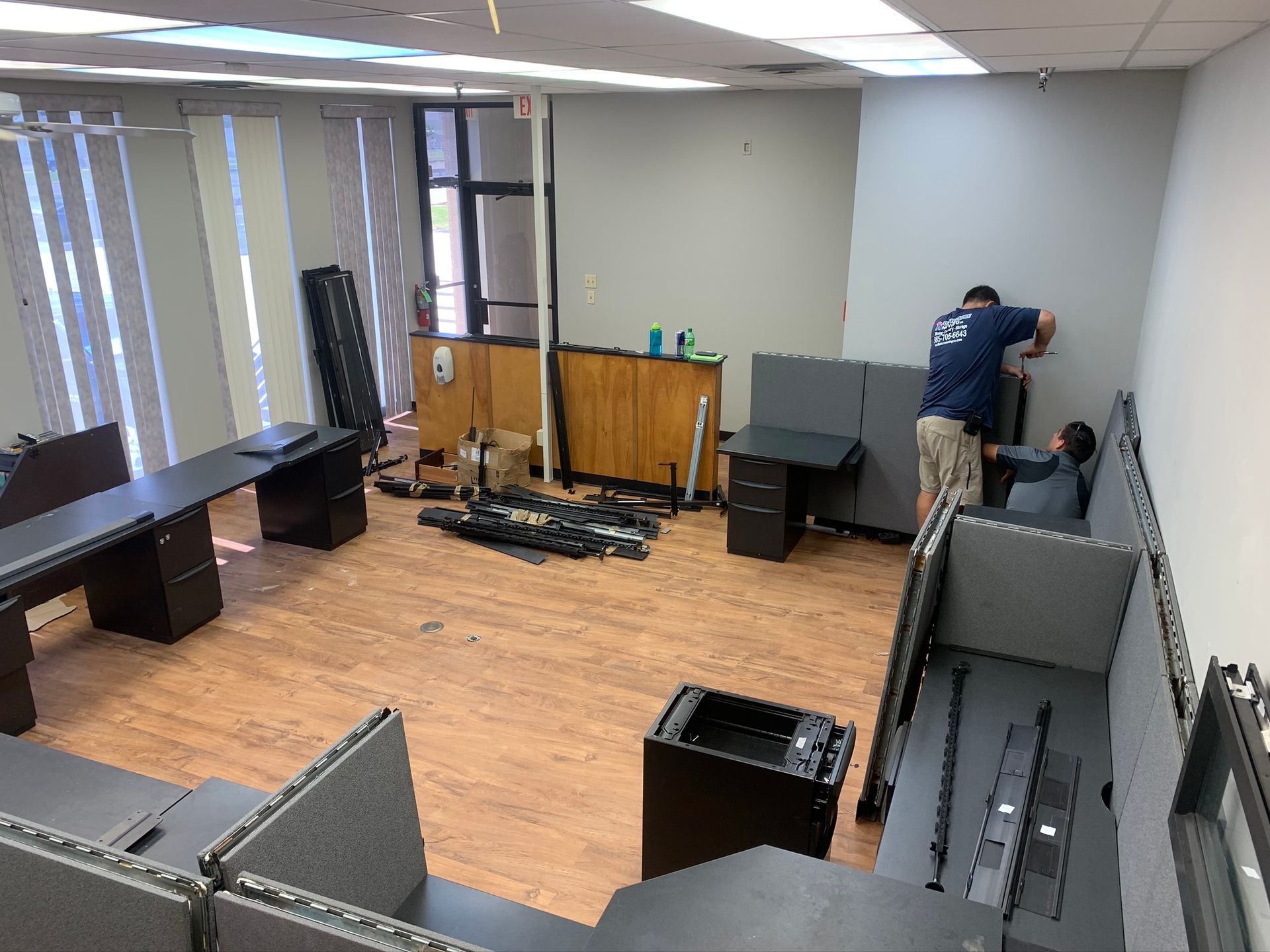 Two people assemble grey modular office cubicles in a room with light wood-look flooring and tall windows.
