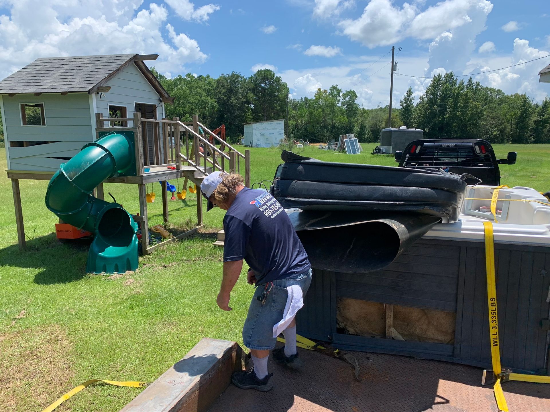 A person works near a hot tub being secured onto a truck bed, next to a backyard playhouse with a green slide.