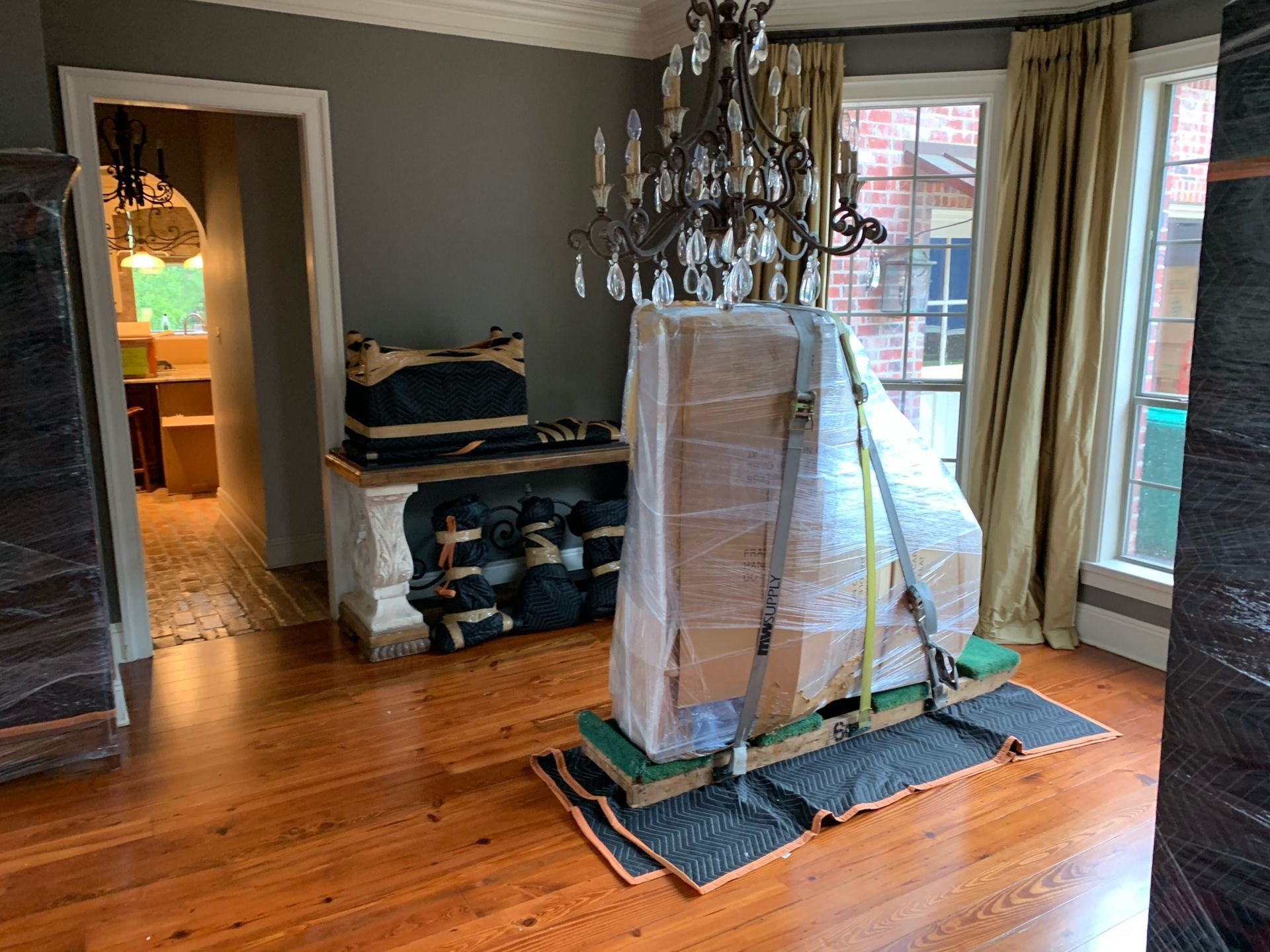 A grand piano, wrapped in plastic and strapped to a moving board, sits in a dining room with wood floors and a chandelier.