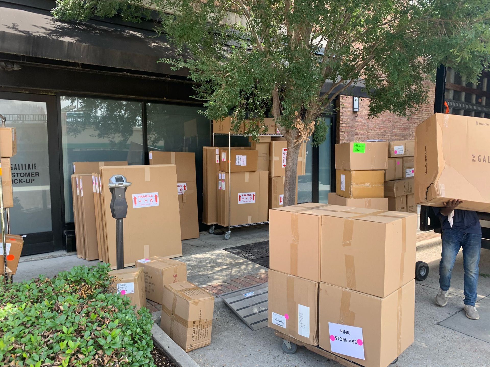 A person carries a large cardboard box toward a building entrance filled with numerous stacked shipping boxes.