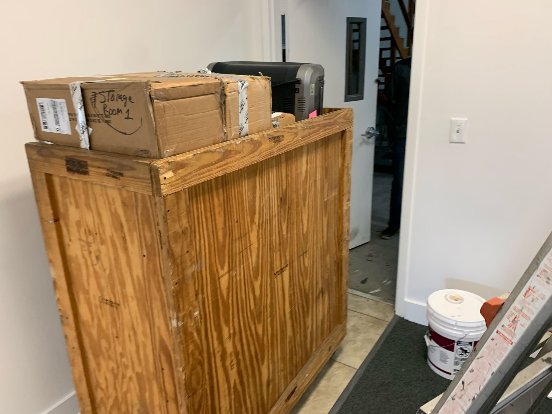 A large wooden crate sits in a hallway, topped with a cardboard box and printer, near a door and a bucket on the floor.