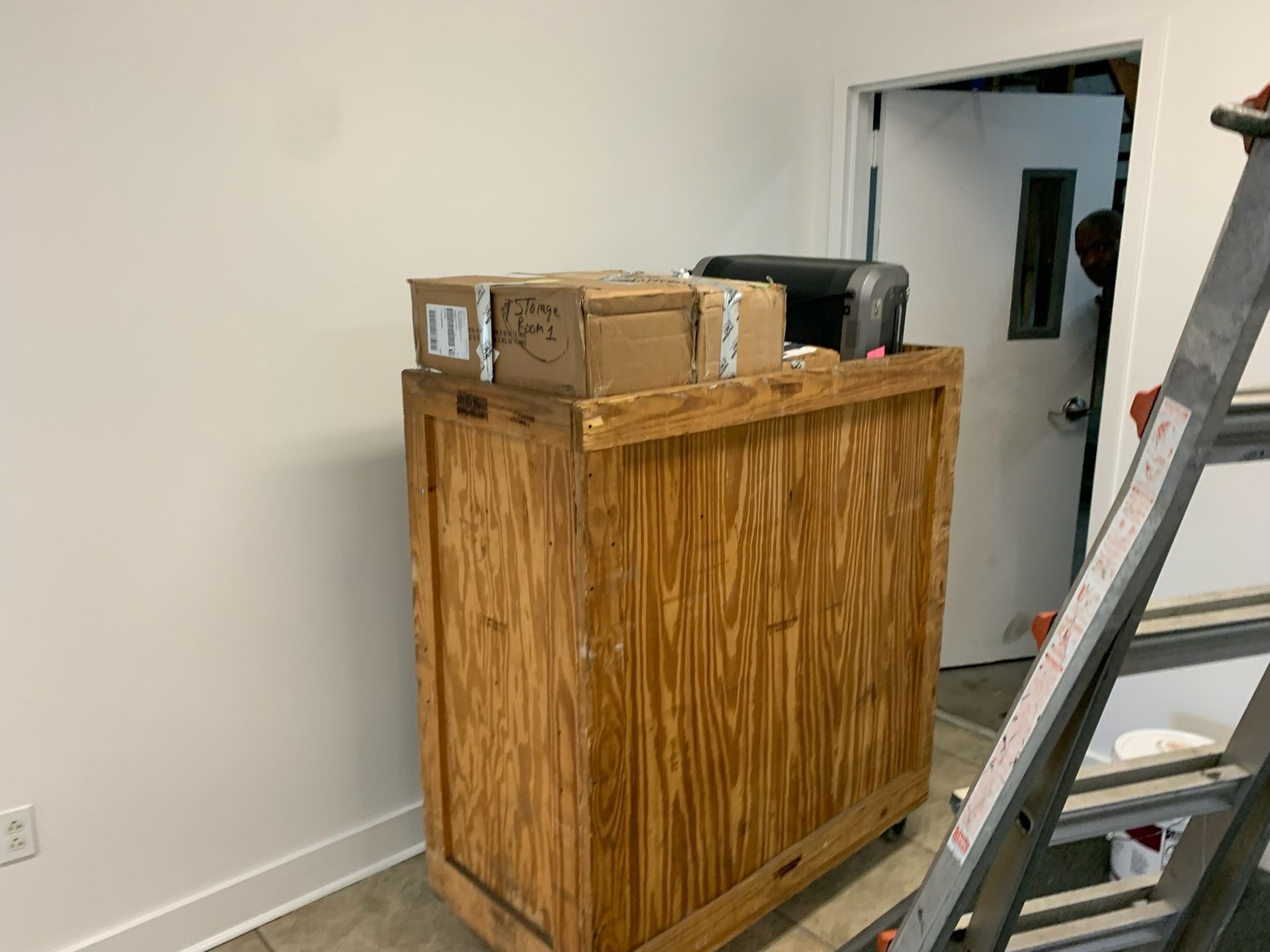 A tall, wooden rolling crate topped with several cardboard boxes, positioned in a room near a doorway and a metal ladder.