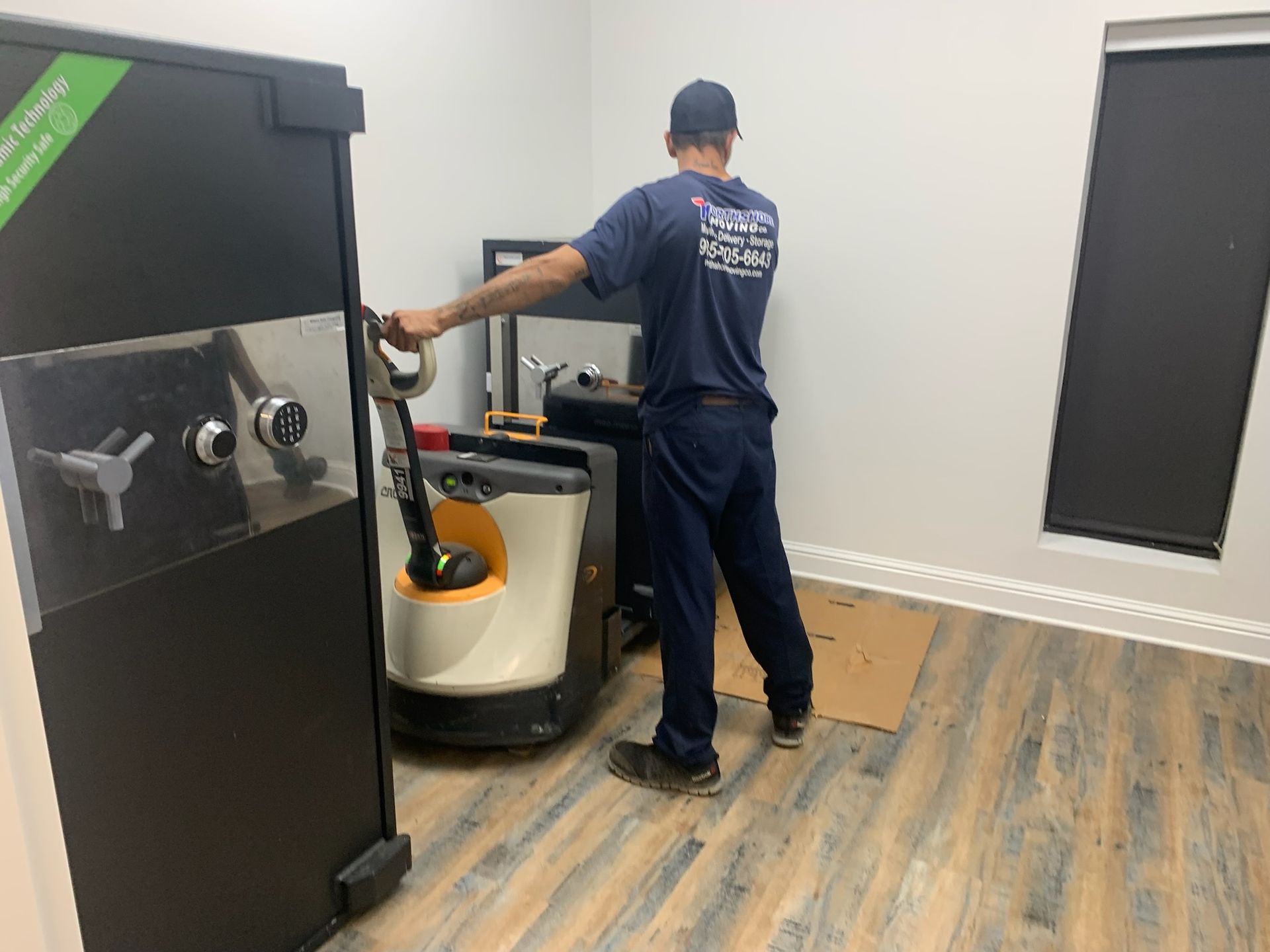 A person in a blue uniform uses a motorized pallet jack to move a large, black safe across a room with wood-look flooring.