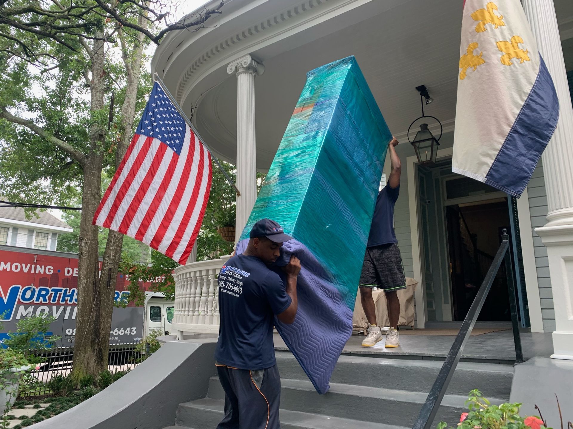 Two movers carry a large, blue-wrapped furniture item up the front steps of a house between two displayed flags.