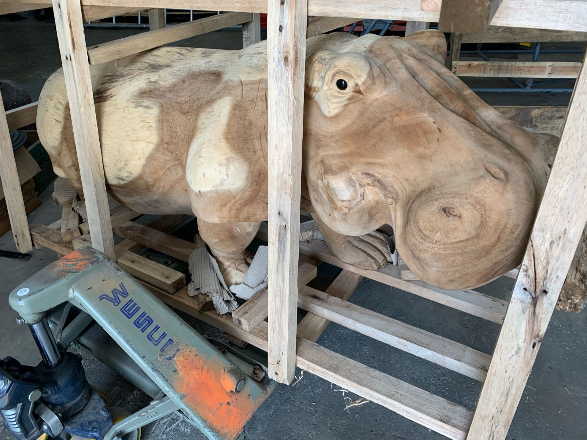 A life-sized wooden carving of a hippopotamus inside a wooden crate, next to an industrial piece of machinery.