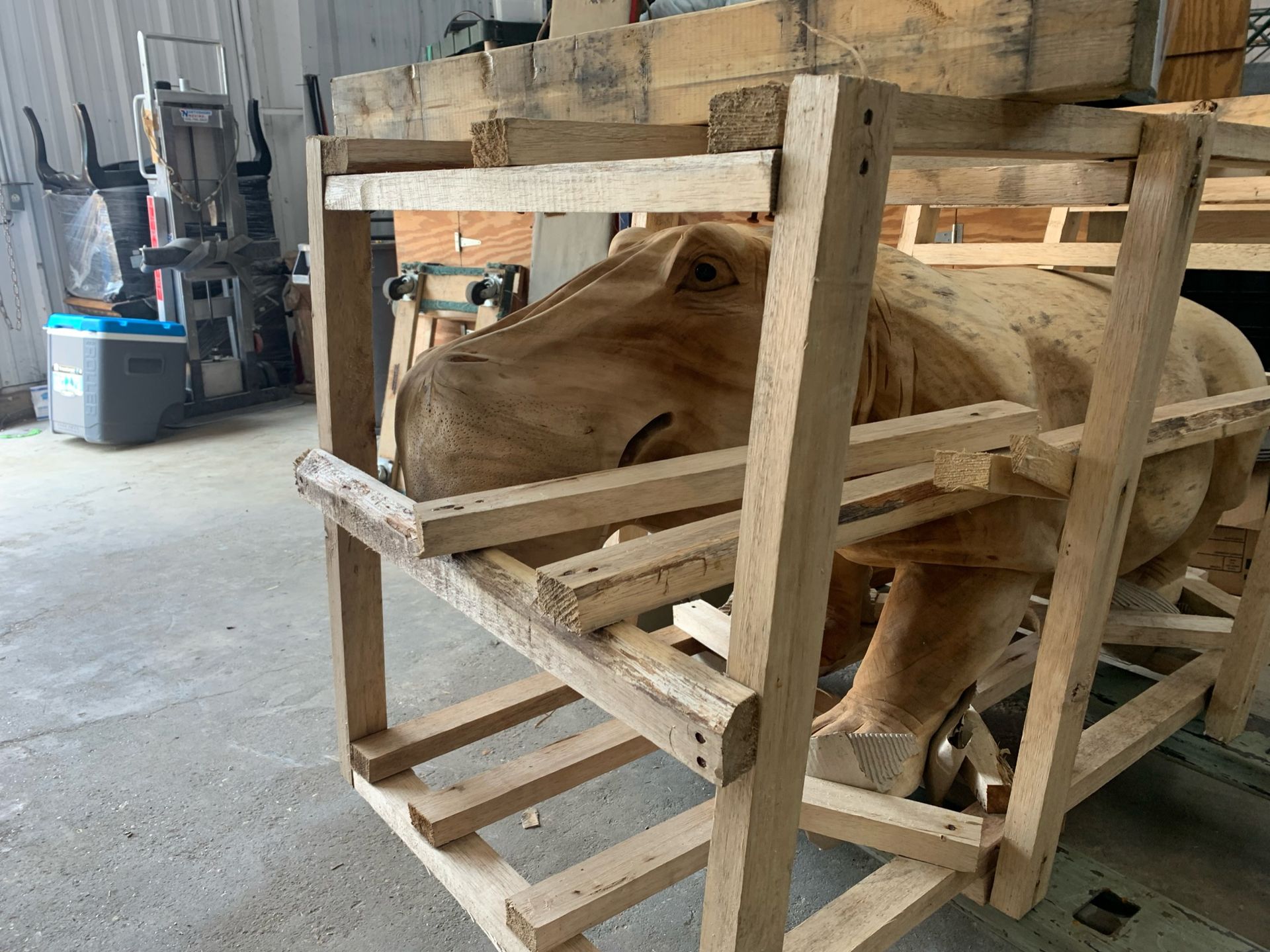 A large wooden hippo sculpture confined inside a wooden crate in a workshop setting.