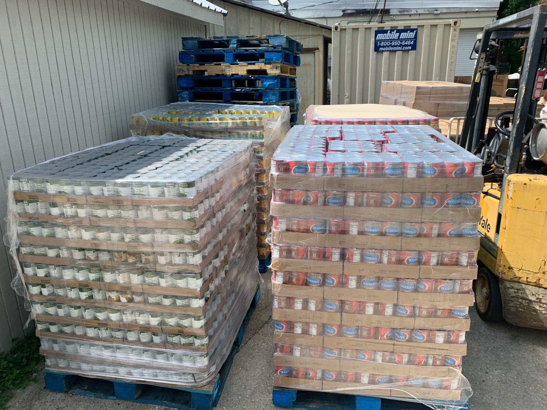 Two pallets of canned goods wrapped in plastic sit outside a warehouse next to a yellow forklift.