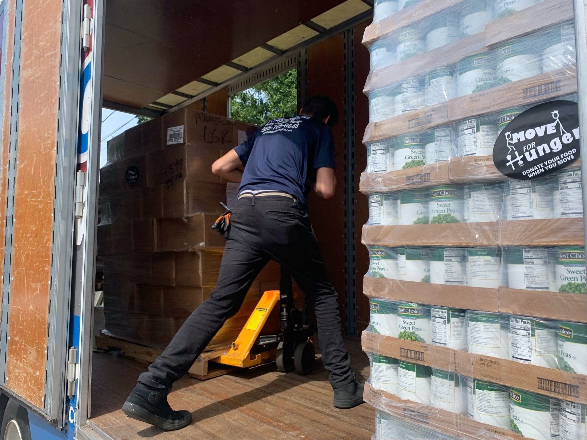 A person wearing a blue shirt and black pants uses a pallet jack to move boxes inside a truck, beside stacks of canned food.