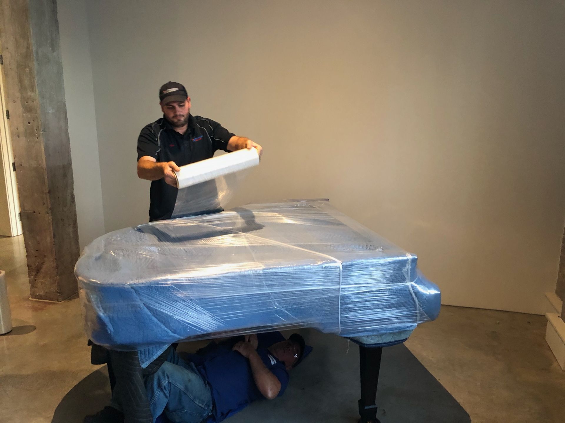 Two movers prepare a grand piano wrapped in protective blue plastic for transport in a room with a concrete floor.