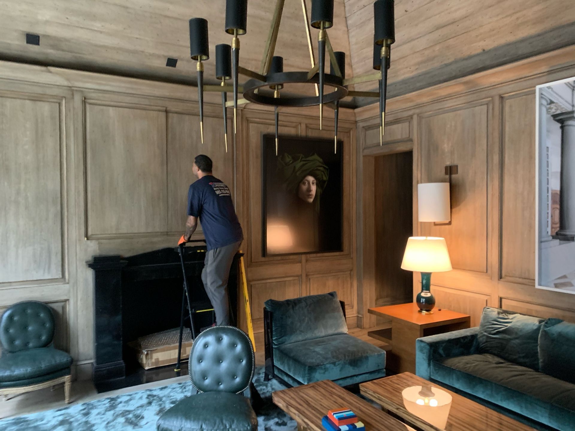 A person stands on a ladder cleaning a black fireplace in a room with wood-paneled walls, a chandelier, and teal furniture.