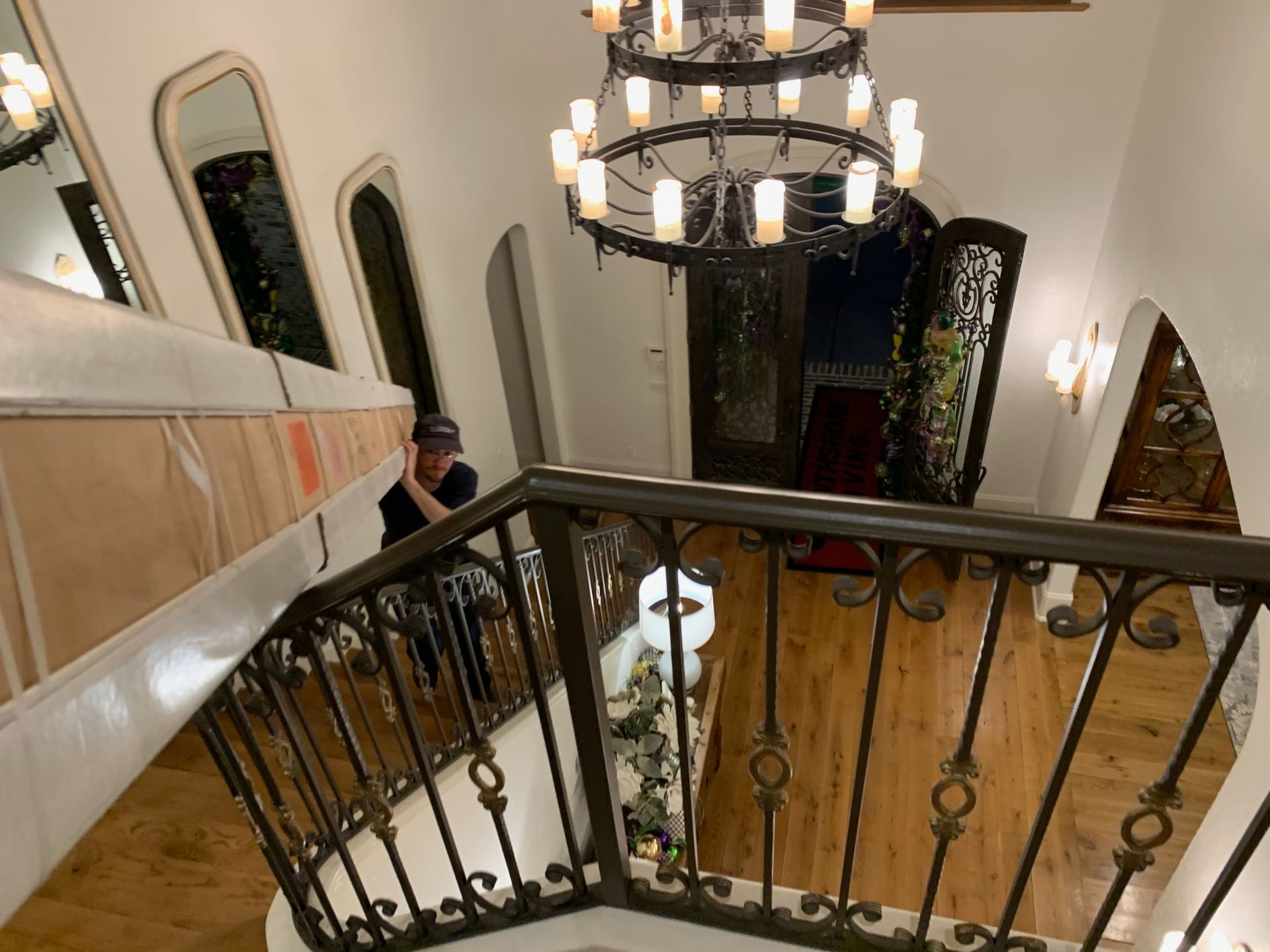 A person carries a large, flat, cardboard-wrapped package down a curved wooden staircase near an ornate chandelier.