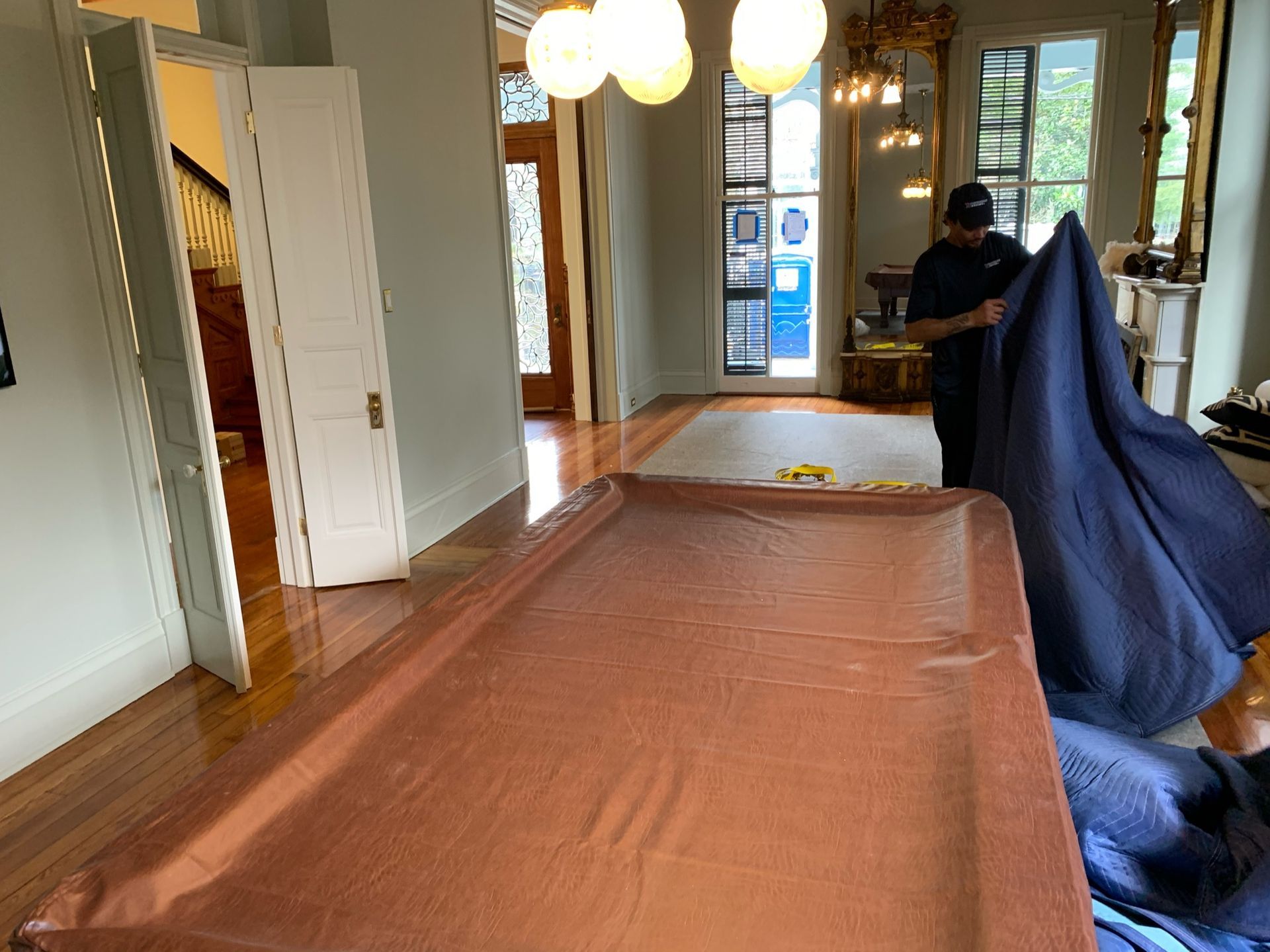 A person draping a protective blue moving blanket over a large pool table in a bright, wood-floored room.