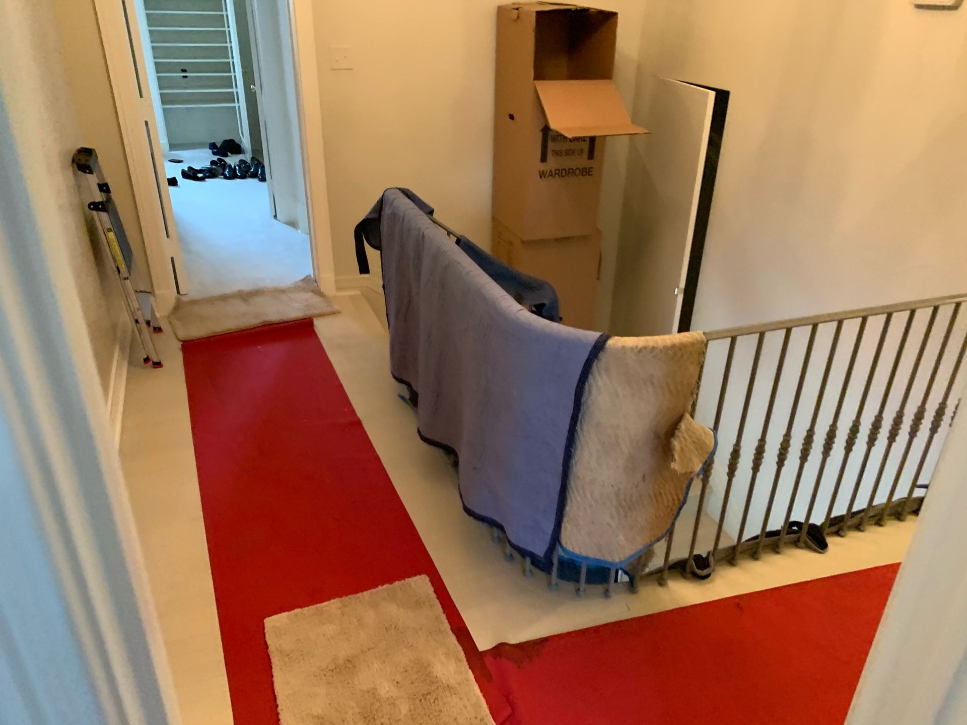 A hallway with red protective floor covering, leading to a staircase, with moving supplies and a blanket-covered item.