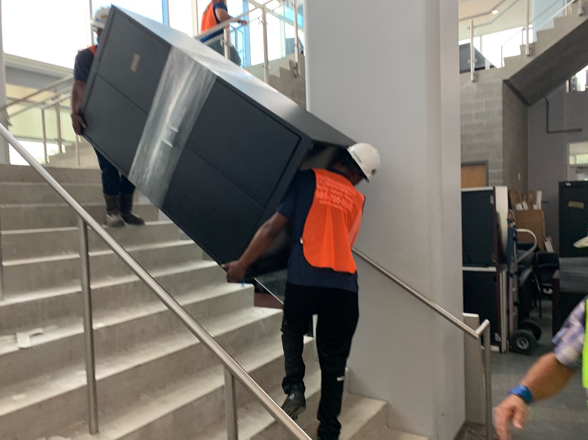 Two workers in safety gear carry a large, dark rectangular cabinet up a concrete staircase inside a building.