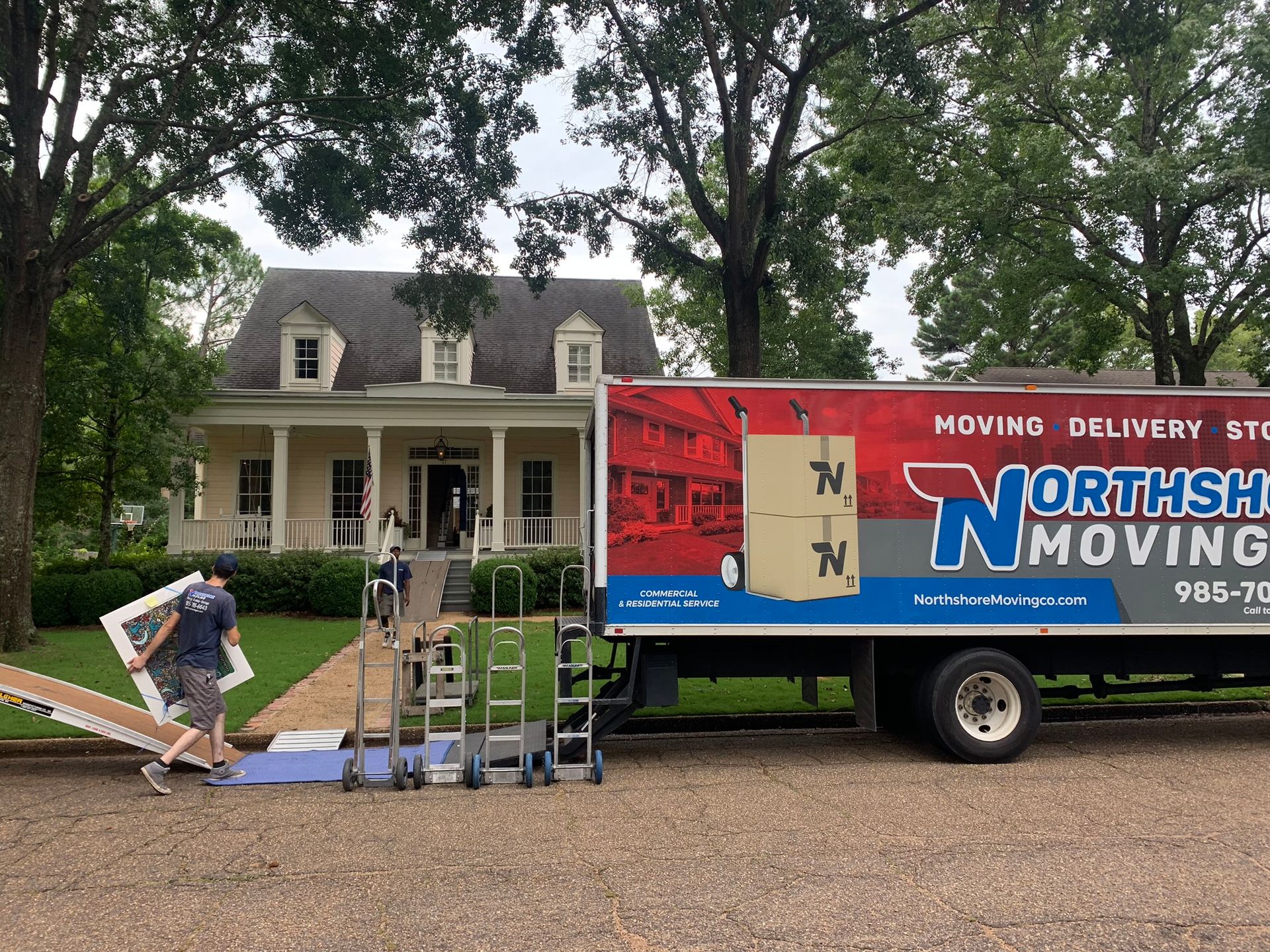 A mover carries a large piece of furniture from a Northshore Moving truck parked in front of a colonial-style house.