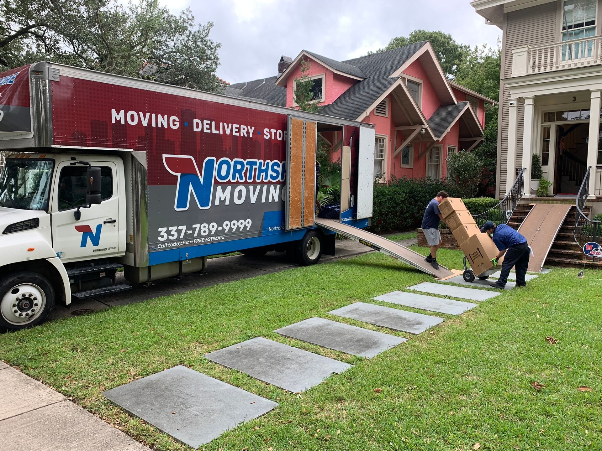 Two workers unload boxes from a Northside Moving truck parked on the grass in front of a house.