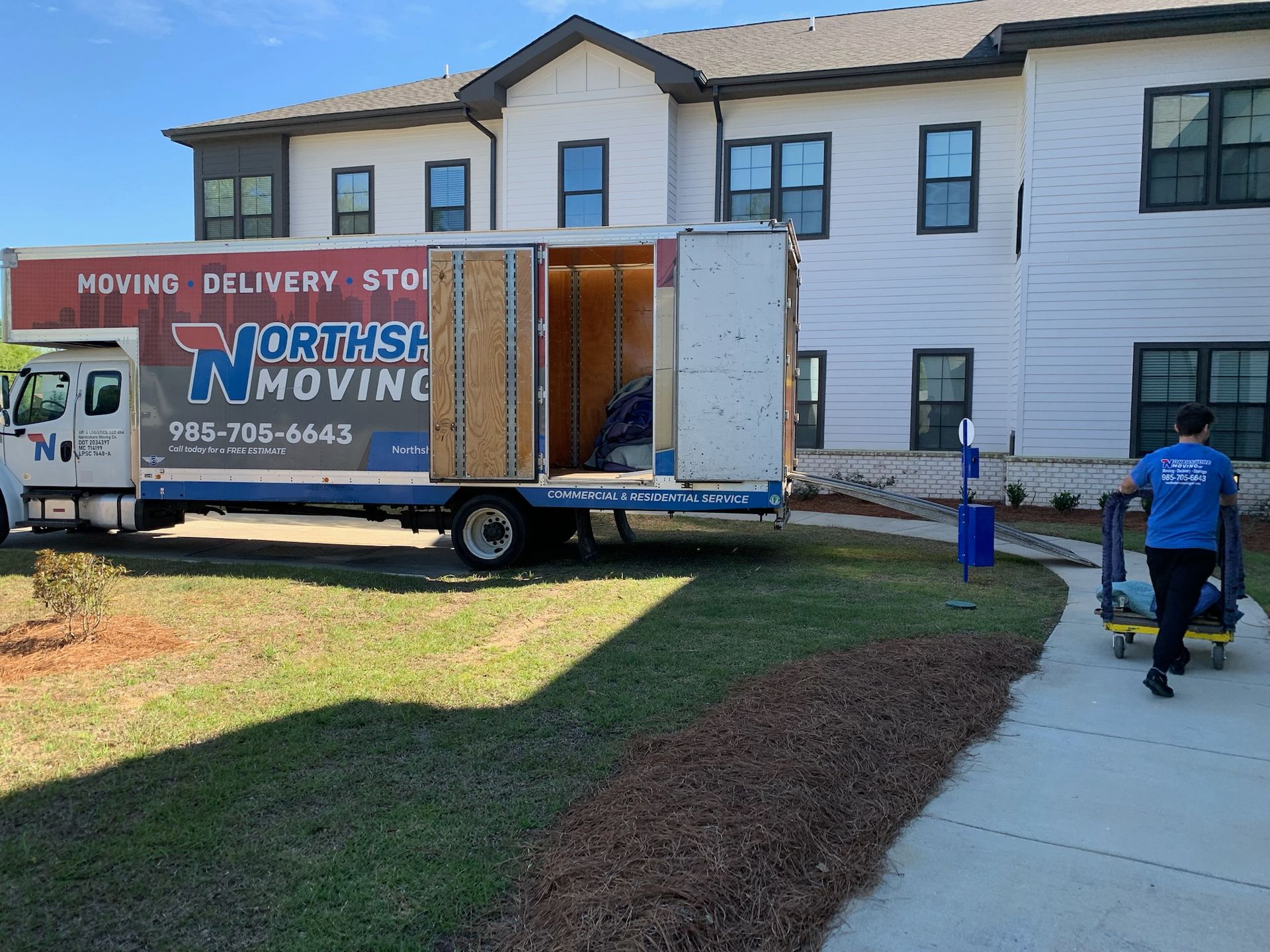 A moving truck parked near a multi-story building as a person in a blue shirt pushes a dolly toward the entrance.