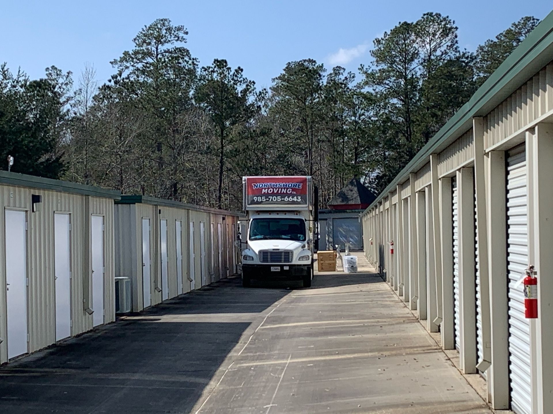 A white moving truck parked in a narrow drive between two rows of beige storage unit doors under a blue sky.