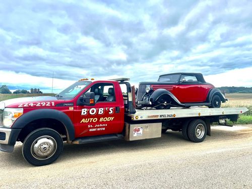 Red tow truck hauling a red vintage convertible on a flatbed under a cloudy sky.