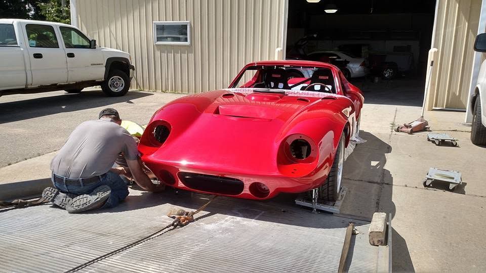 Person kneels by a red sports car outside a garage, polishing its front end.