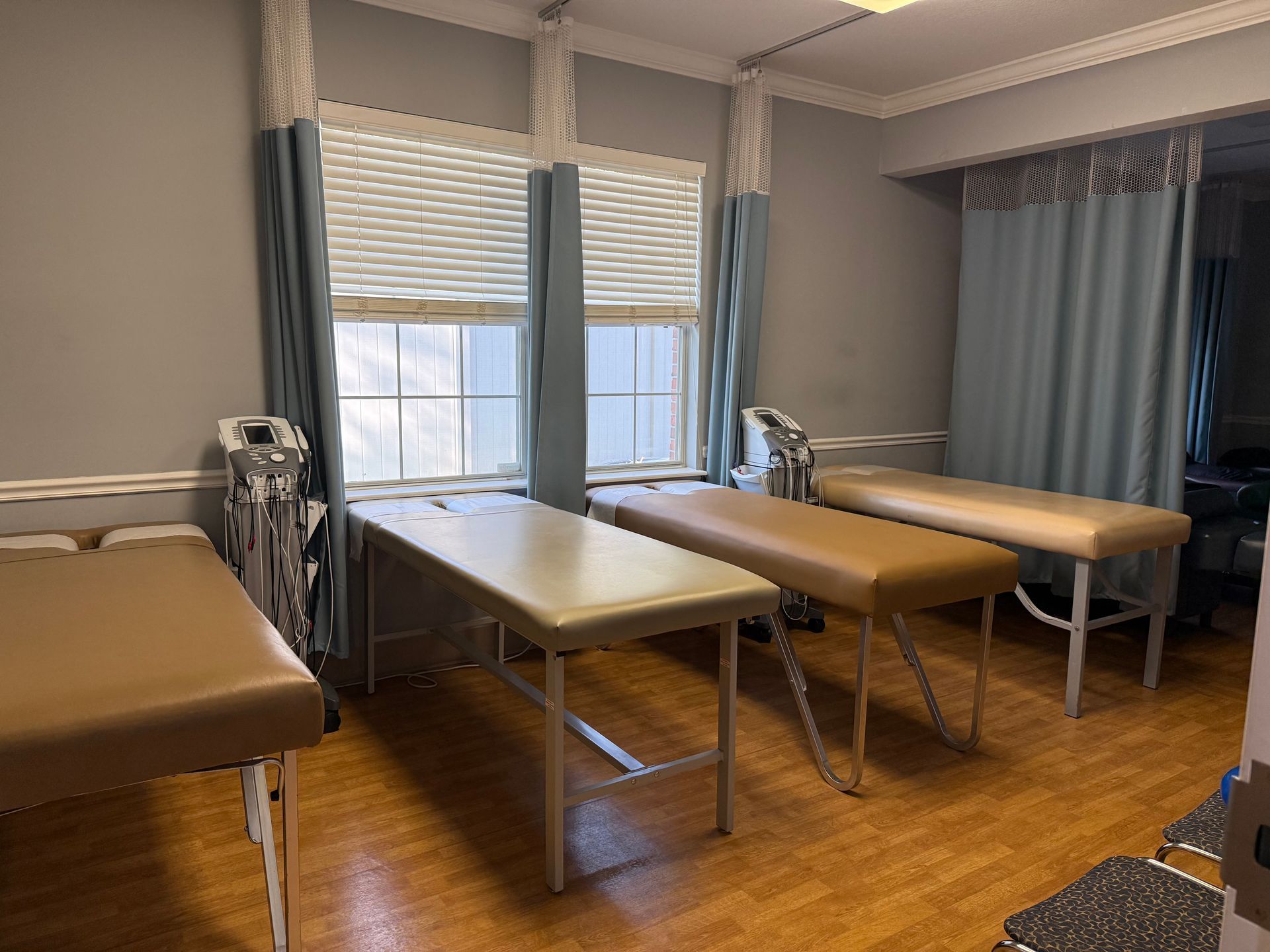 Empty physical therapy room with three treatment tables, window blinds, and blue curtains