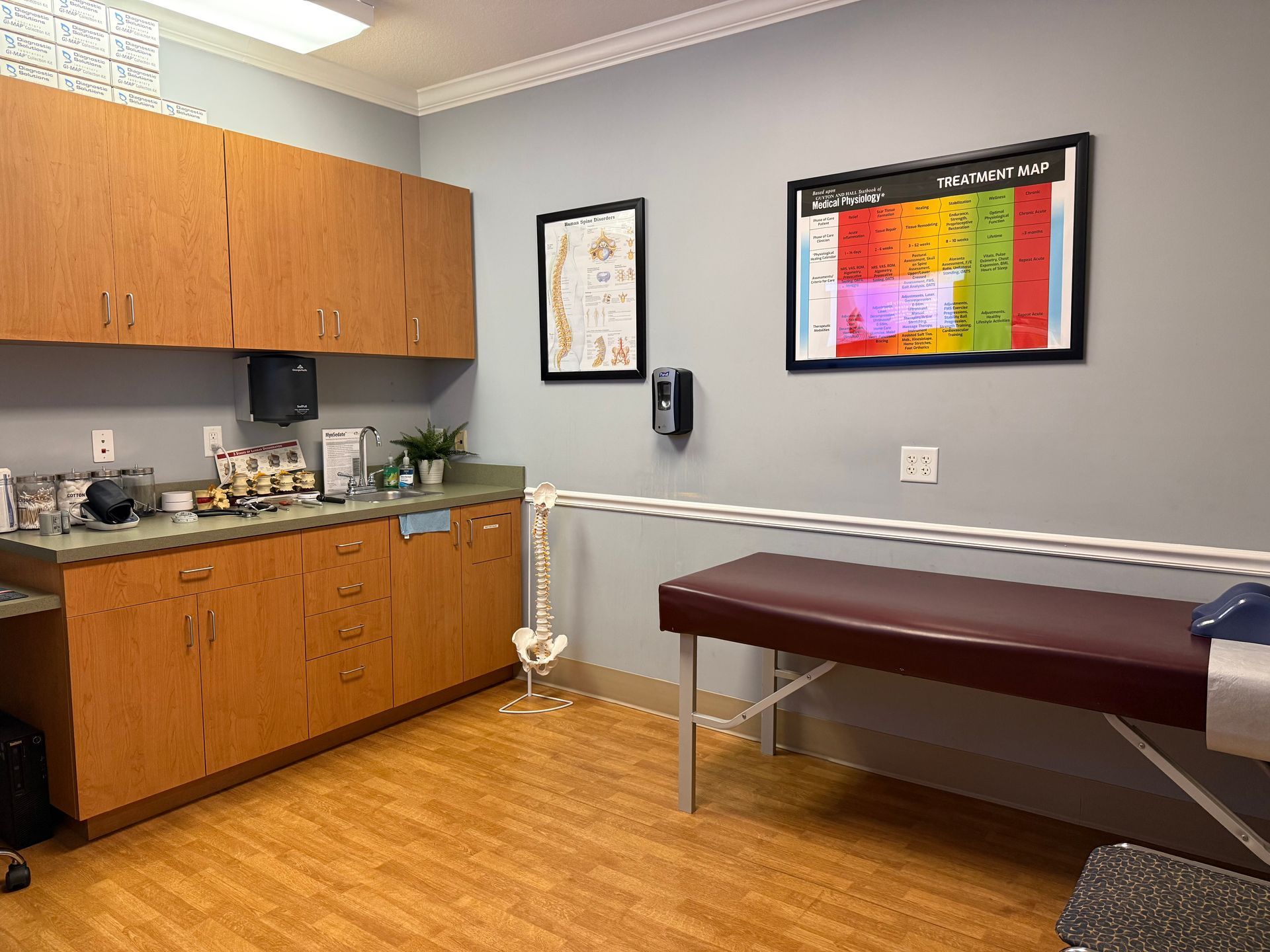 Clinic exam room with cabinets, sink, exam table, and colorful wall chart on gray walls