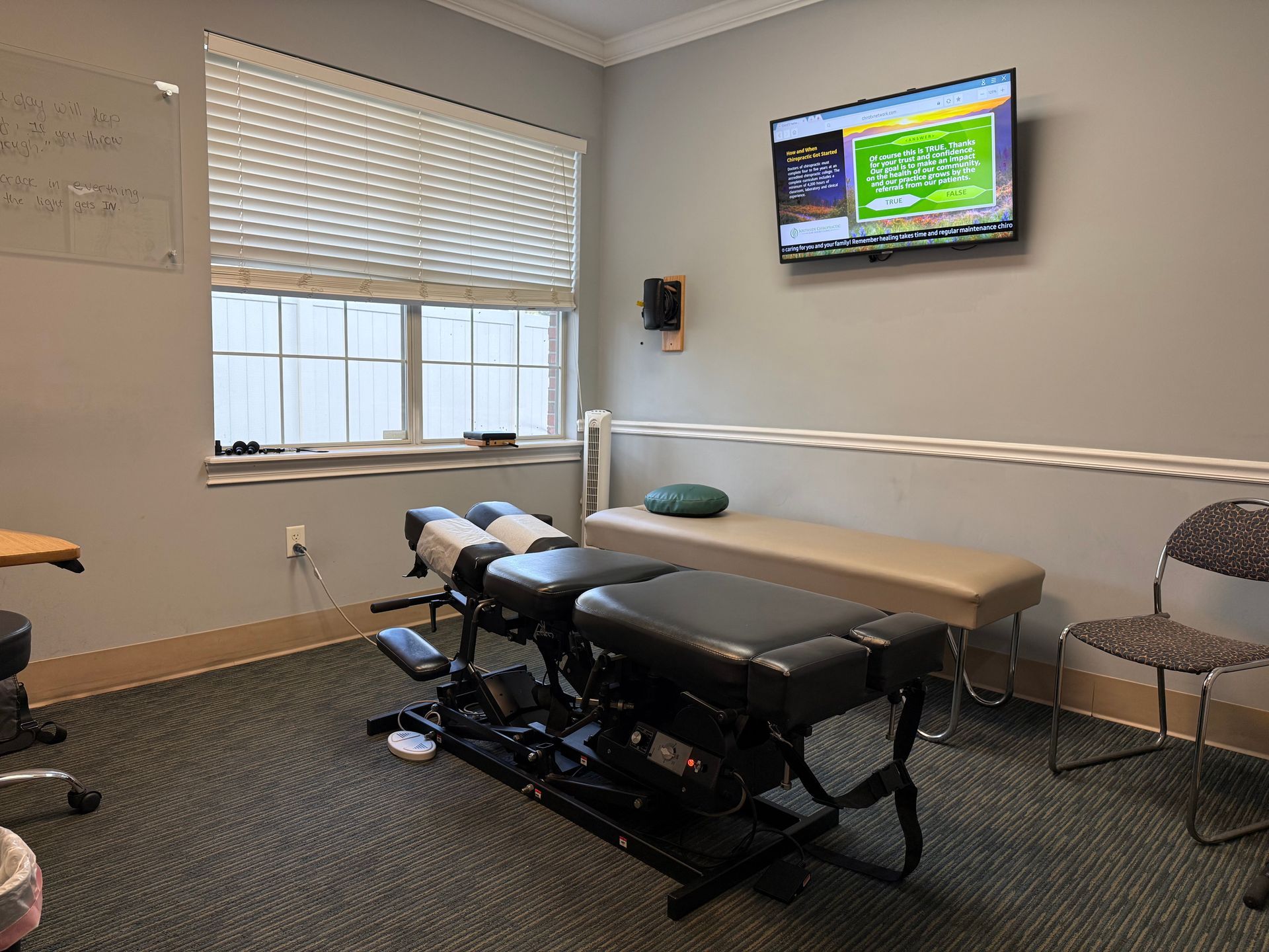 Clinic treatment room with exam table, wall-mounted TV, window blinds, and chairs