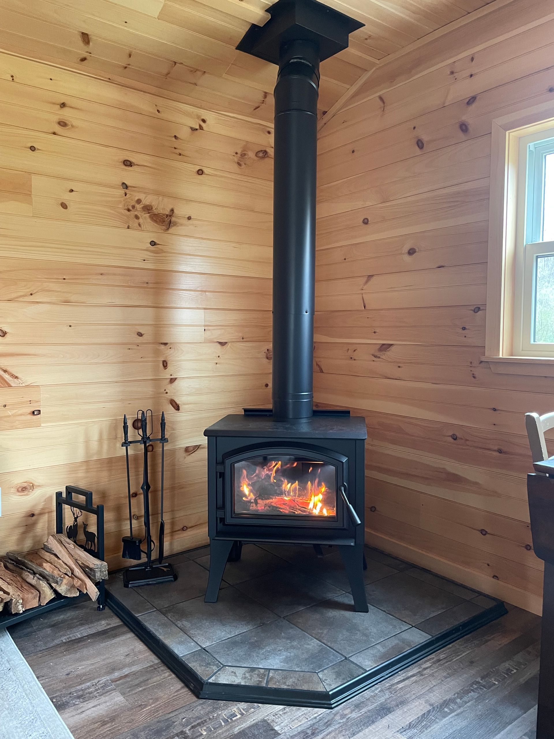 Wood stove burning in a cozy pine-paneled cabin corner, with firewood and tools nearby.