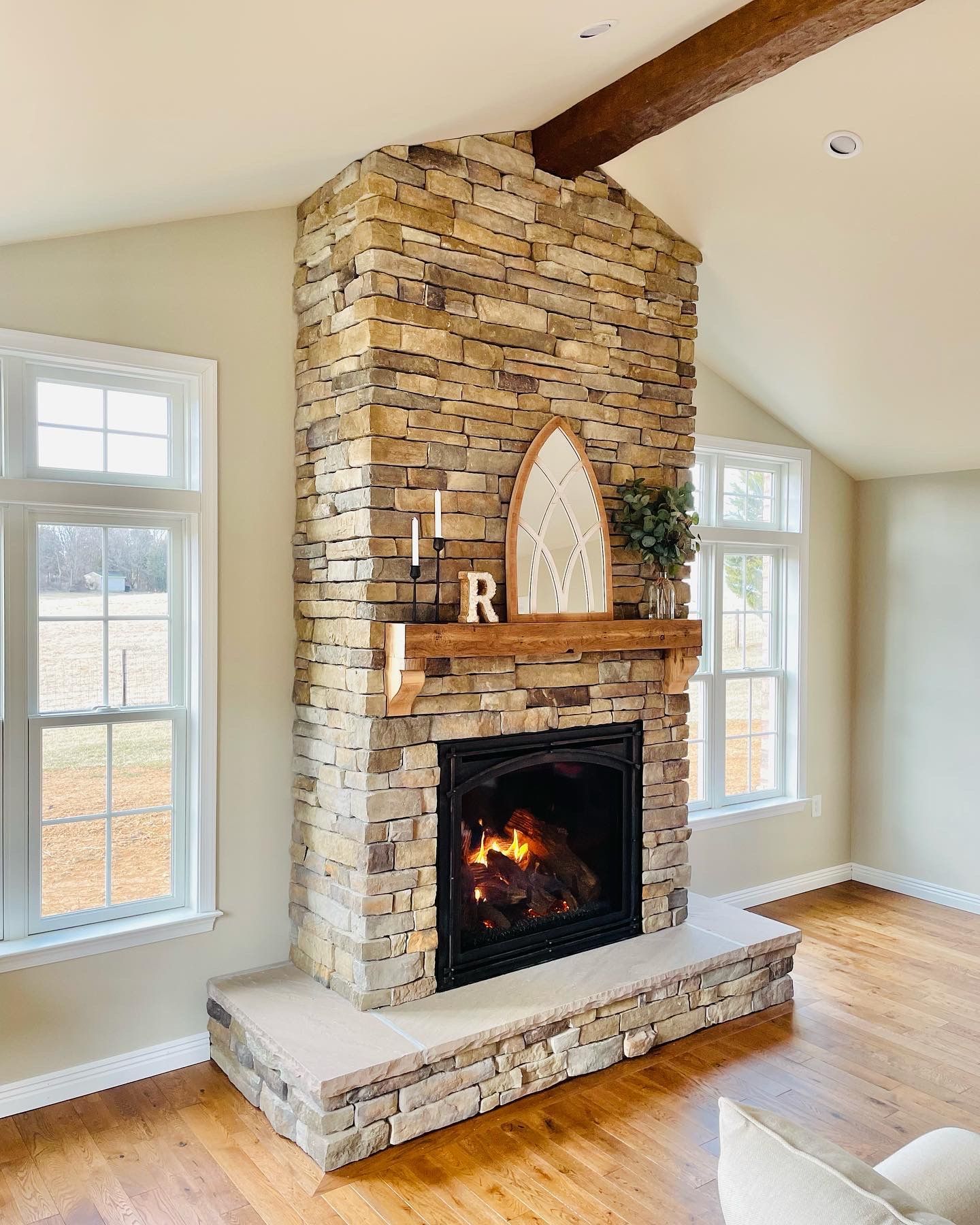 Stone fireplace with lit fire and mantel in a bright living room with large windows