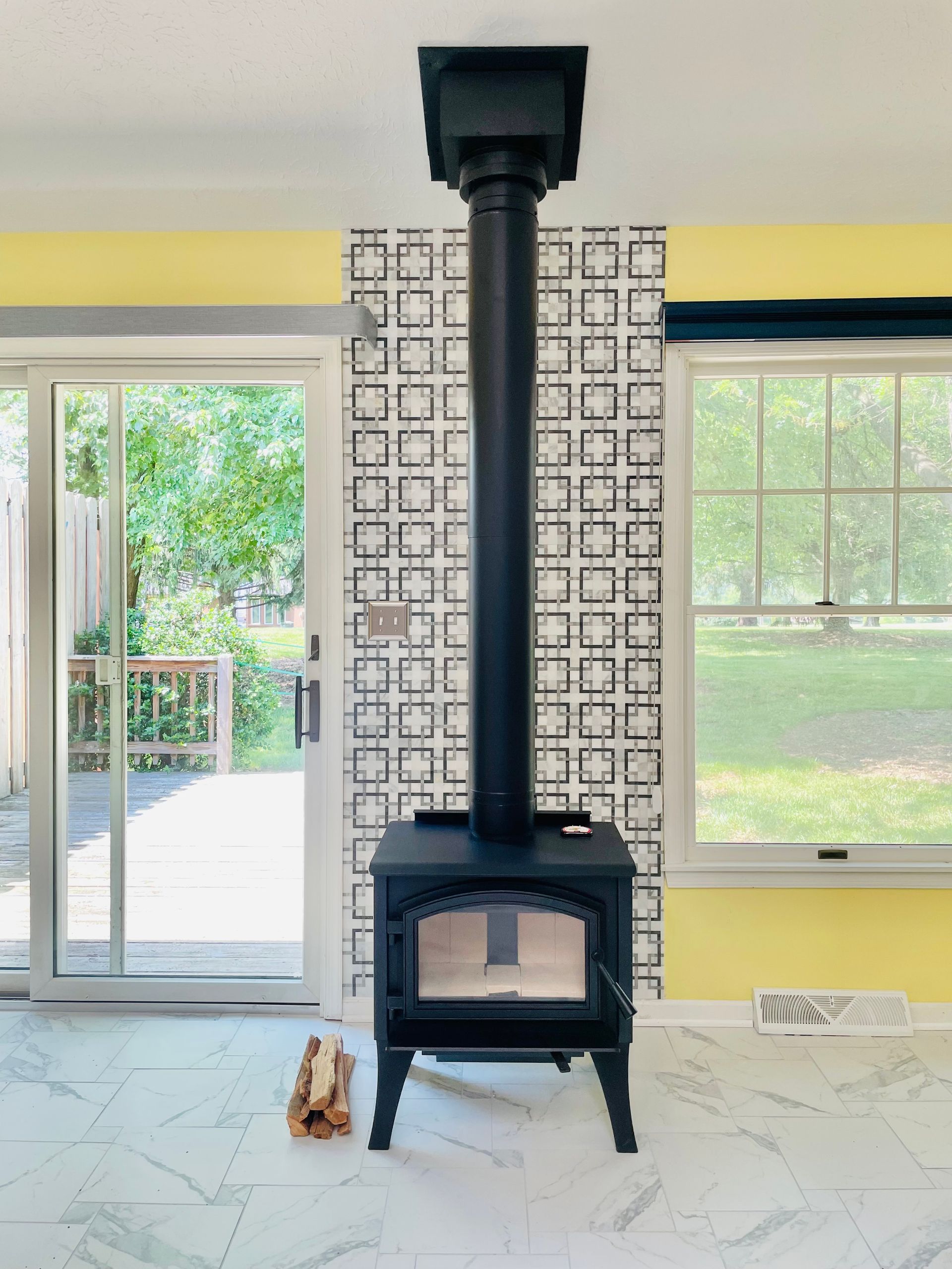 Black wood stove with tall chimney in a bright room with large windows and patterned wall paper