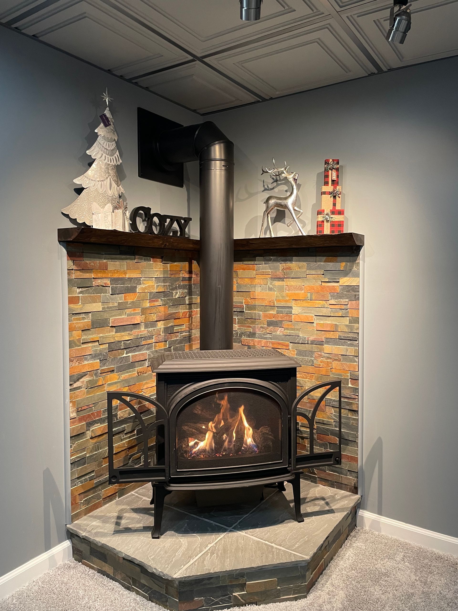 Wood-burning stove in a tiled corner hearth with a black pipe, festive decor, and white tree accents