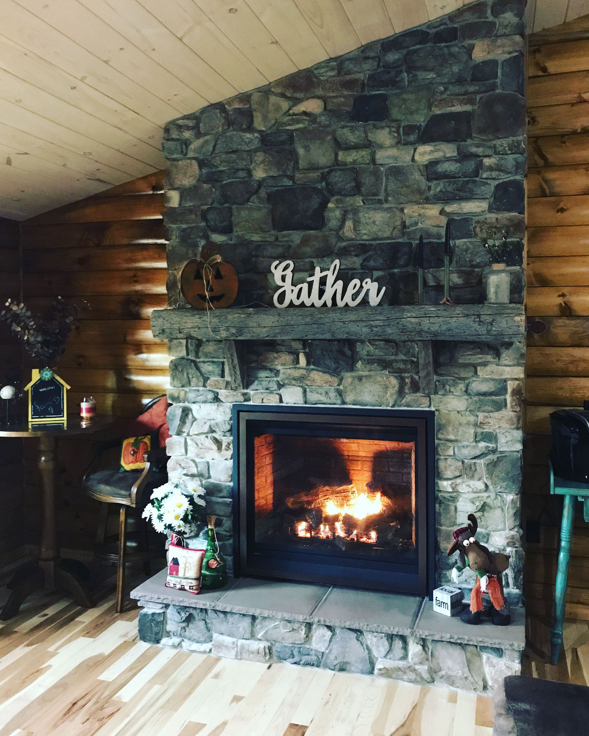 Rustic stone fireplace with a lit fire and “gather” sign in a log cabin living room