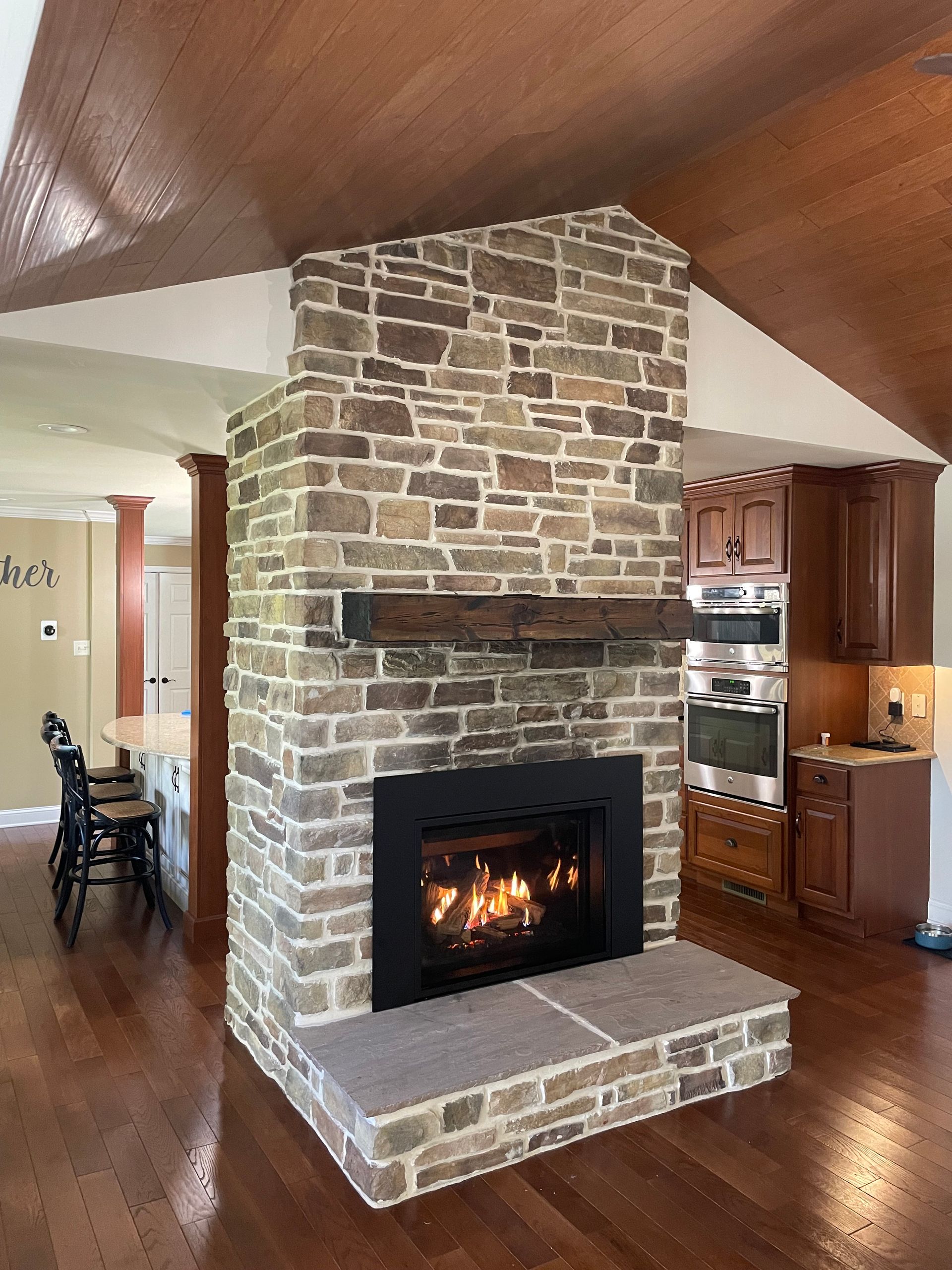 Stone fireplace with lit fire in a wood-floored living room, flanked by a dining area and kitchen.