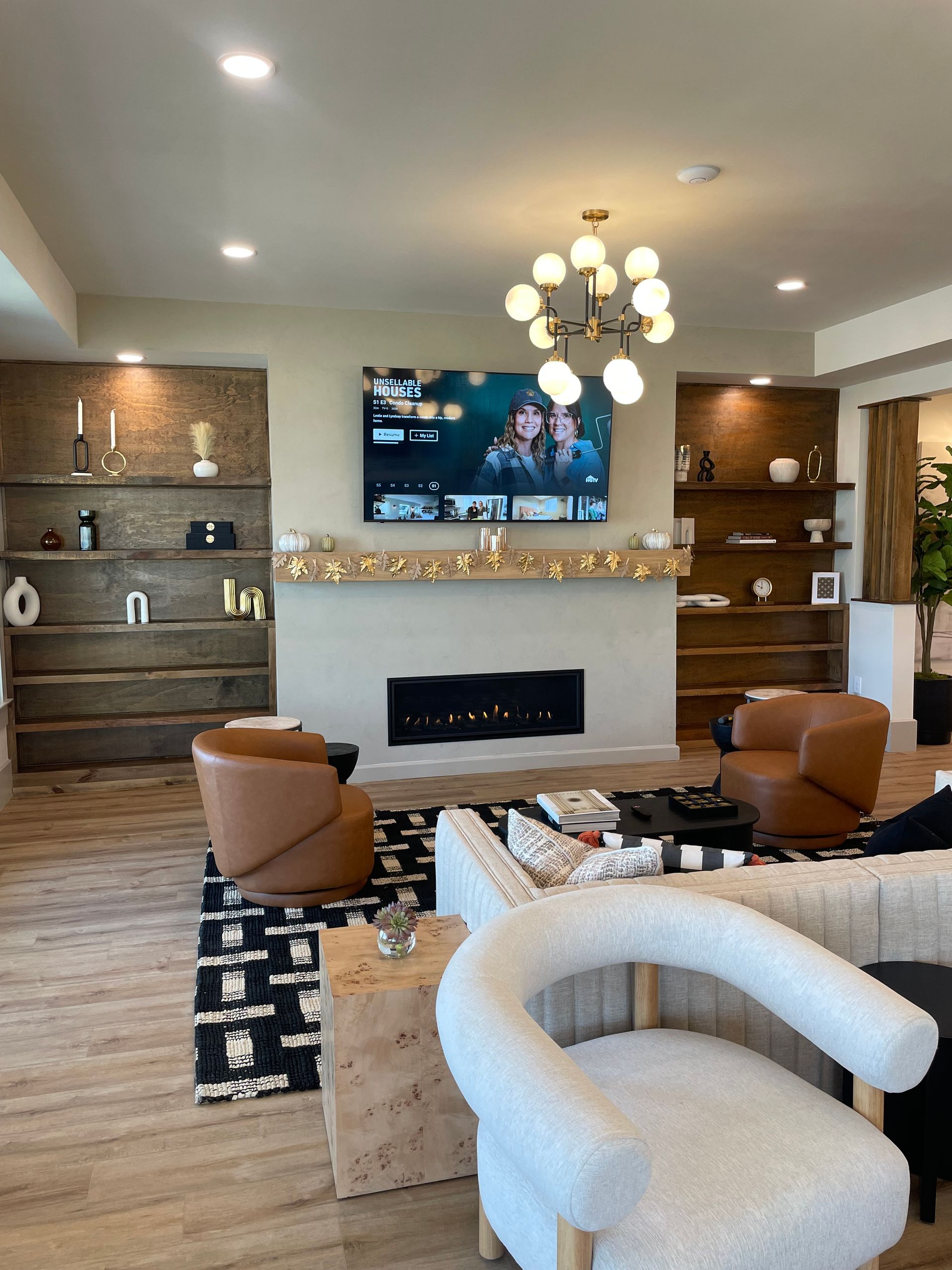 Modern living room with fireplace, wall-mounted TV, chandelier, and tan chairs on a black-and-white rug