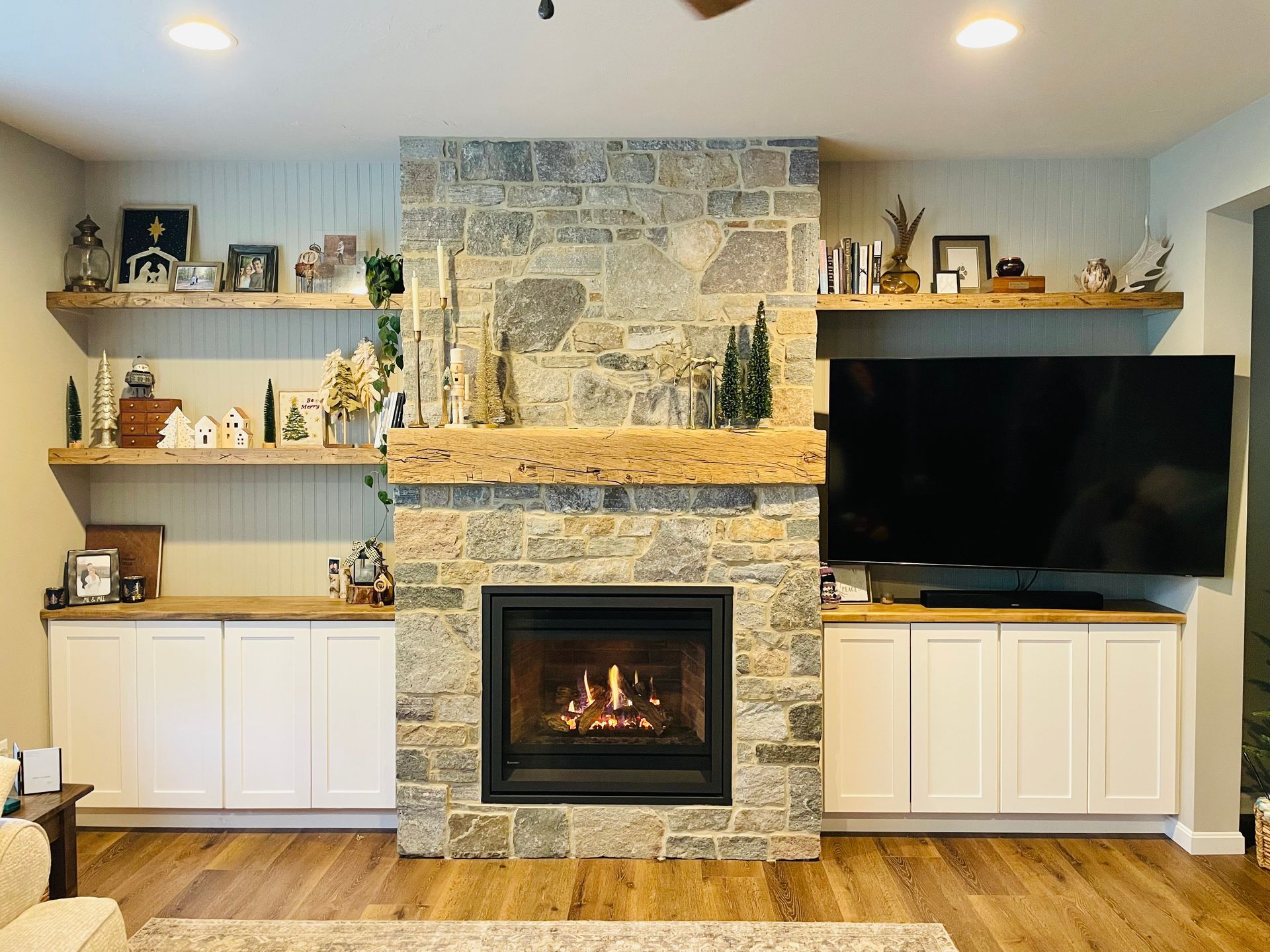 Living room with stone fireplace, TV, built-in shelves, and white cabinets on wood floor