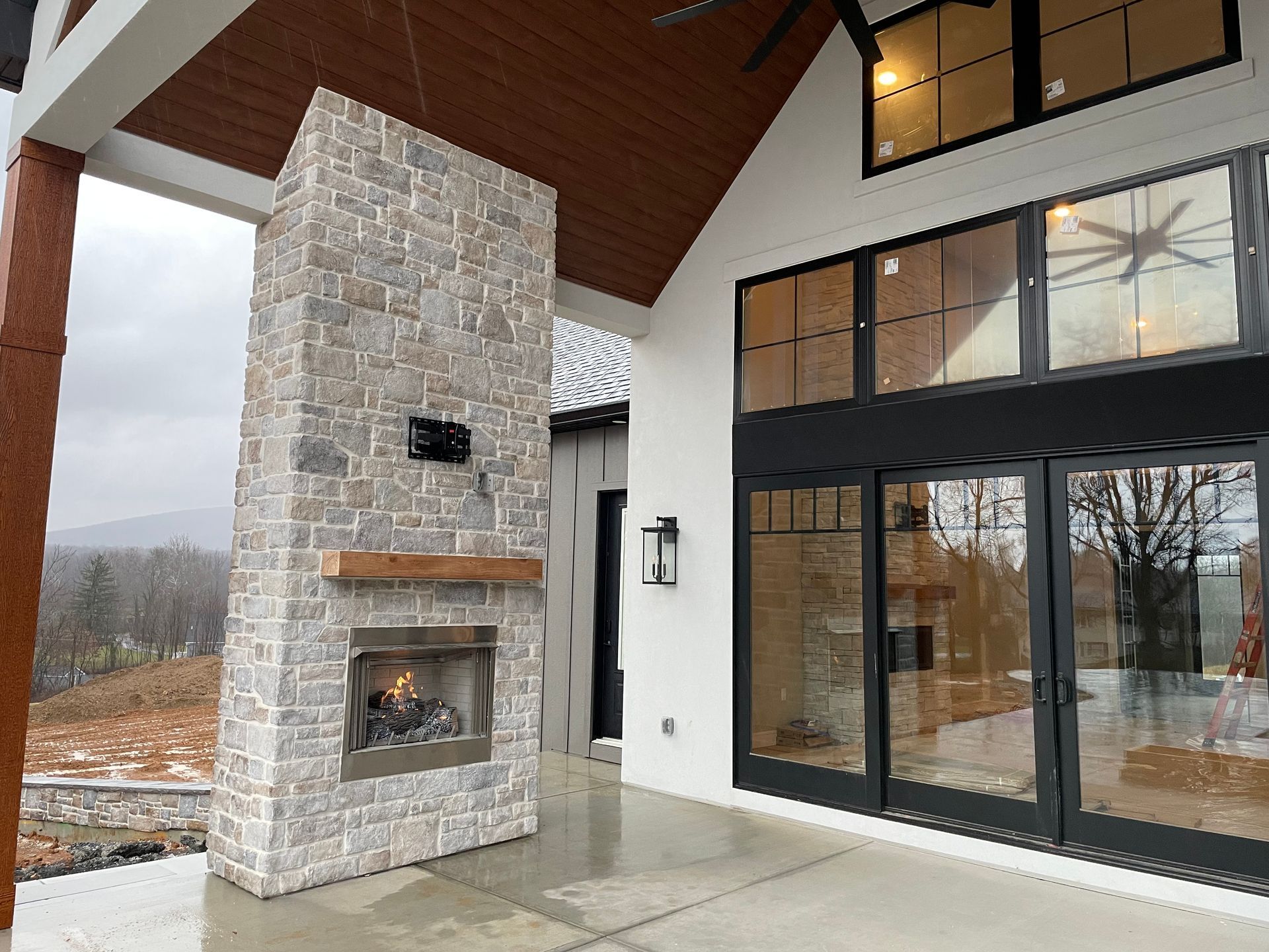 Covered patio with stone fireplace, wood ceiling, and black-framed glass doors overlooking a wet outdoor view