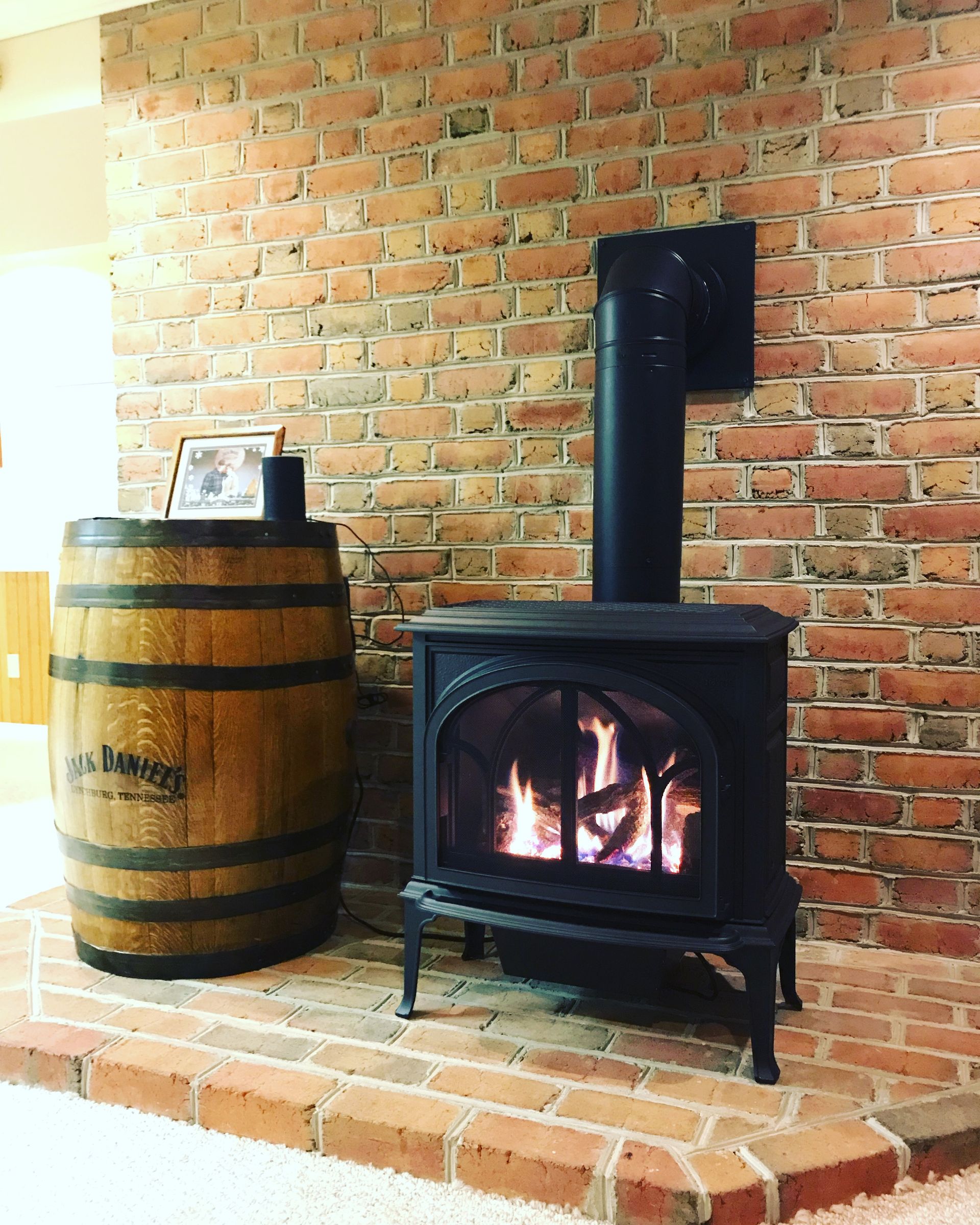 Wood-burning stove with a fire, set against a brick wall beside a wooden barrel.