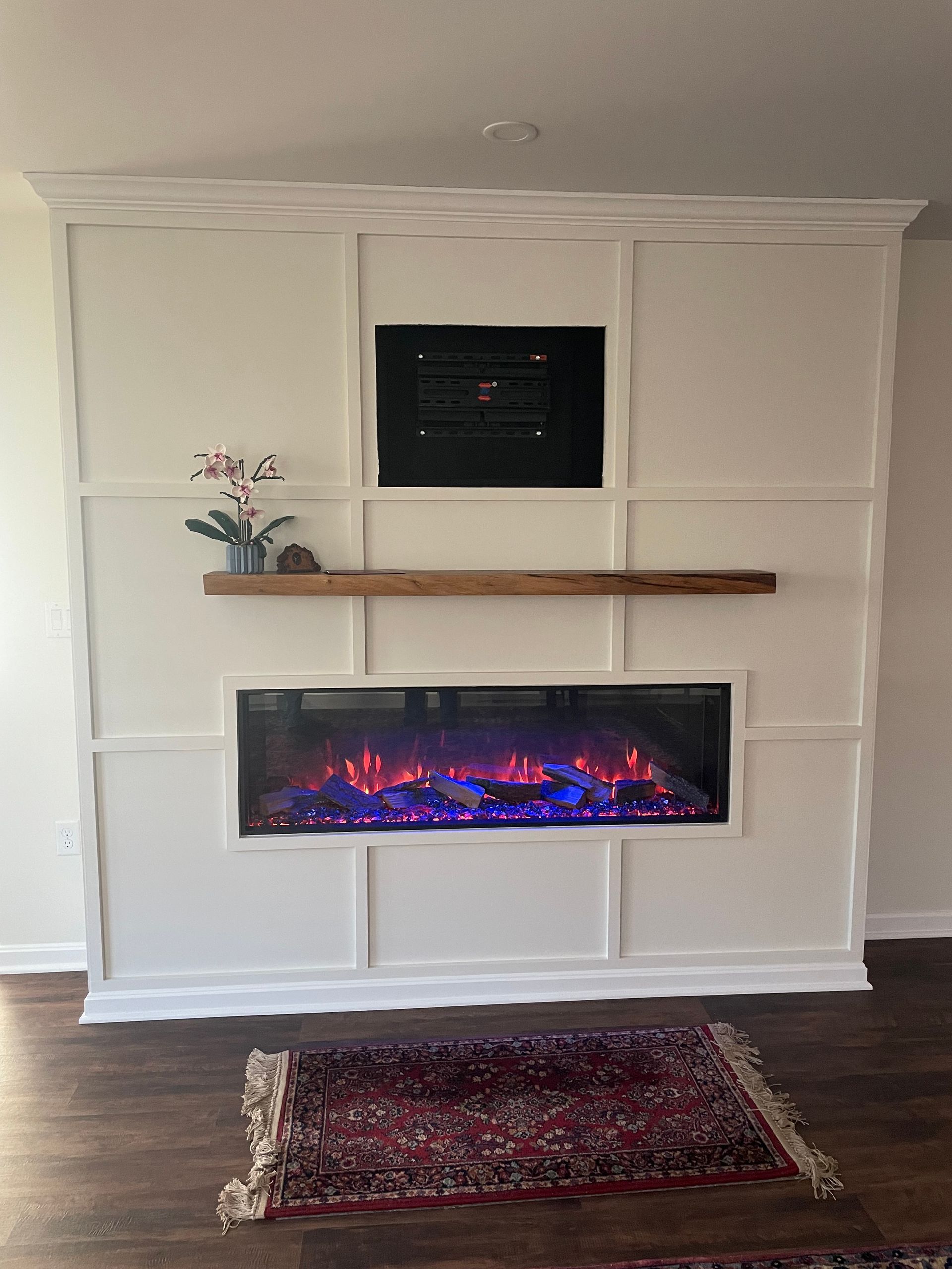 White living room wall with built-in electric fireplace, dark TV above, wood mantel, and patterned rug.